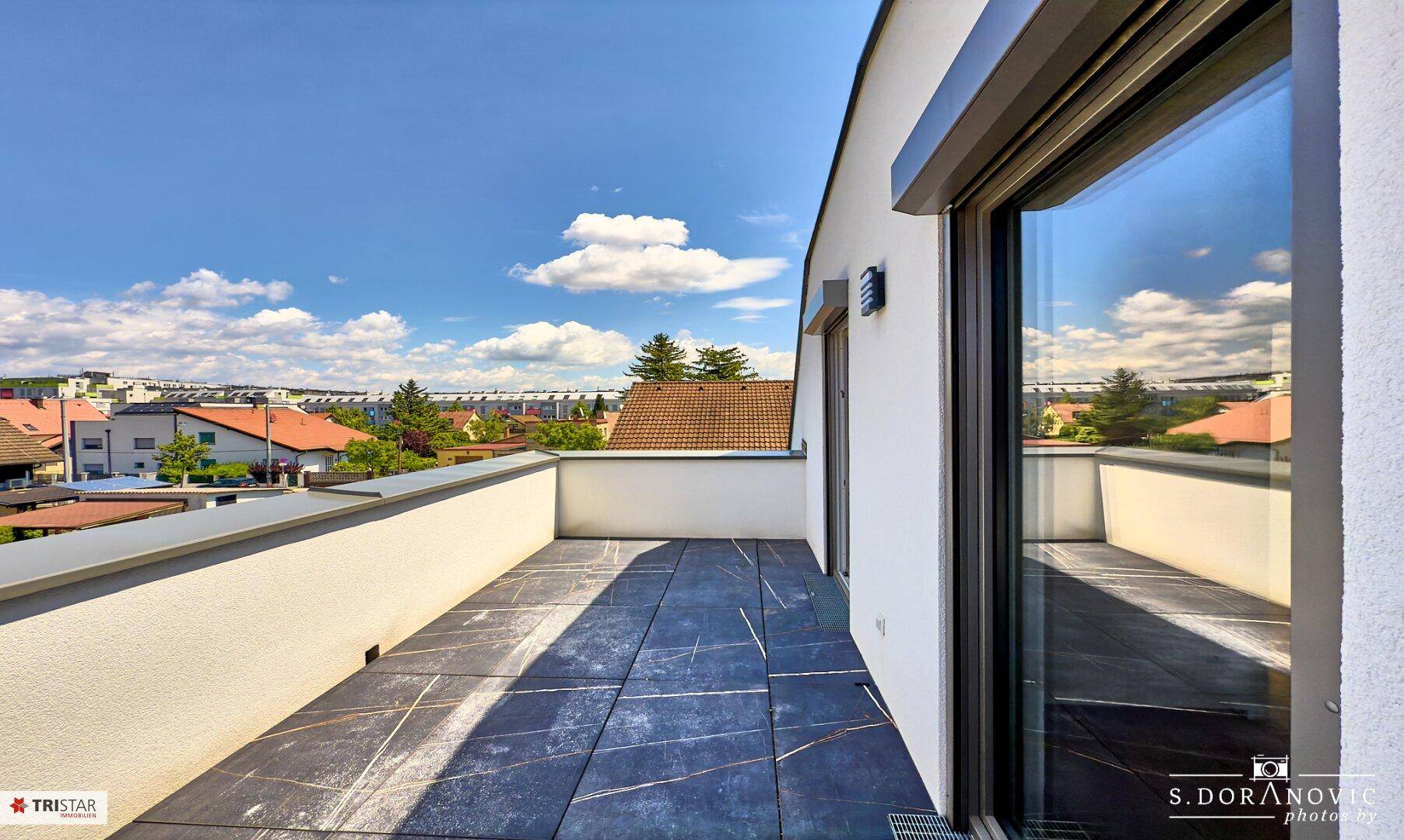 Geräumige Dachterrasse mit modernen Fliesen und großen Fenstertüren unter blauem Himmel.