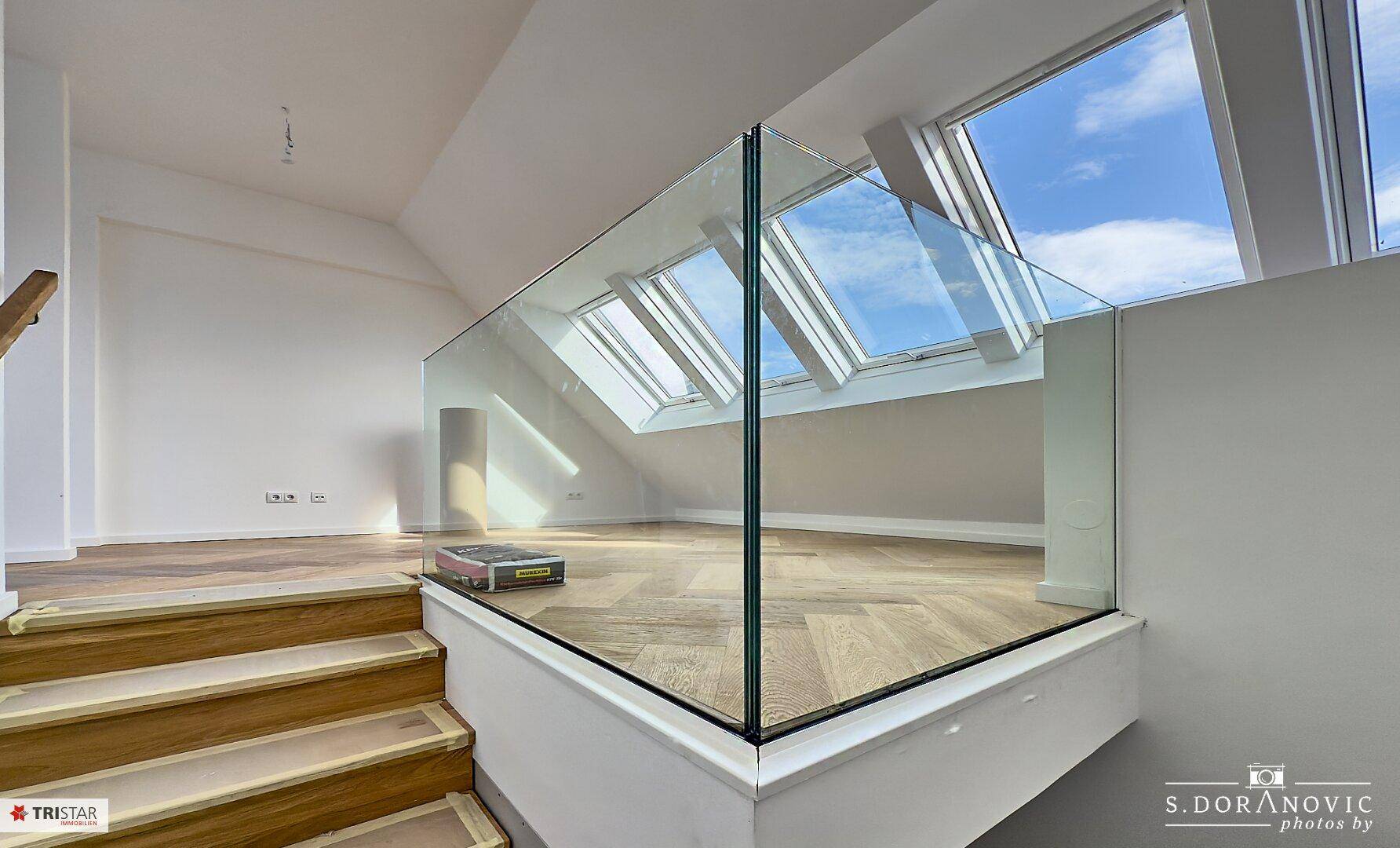 Helles Dachgeschoss mit Holztreppe, Glasgeländer und mehreren Dachfenstern, die viel Tageslicht hereinlassen.