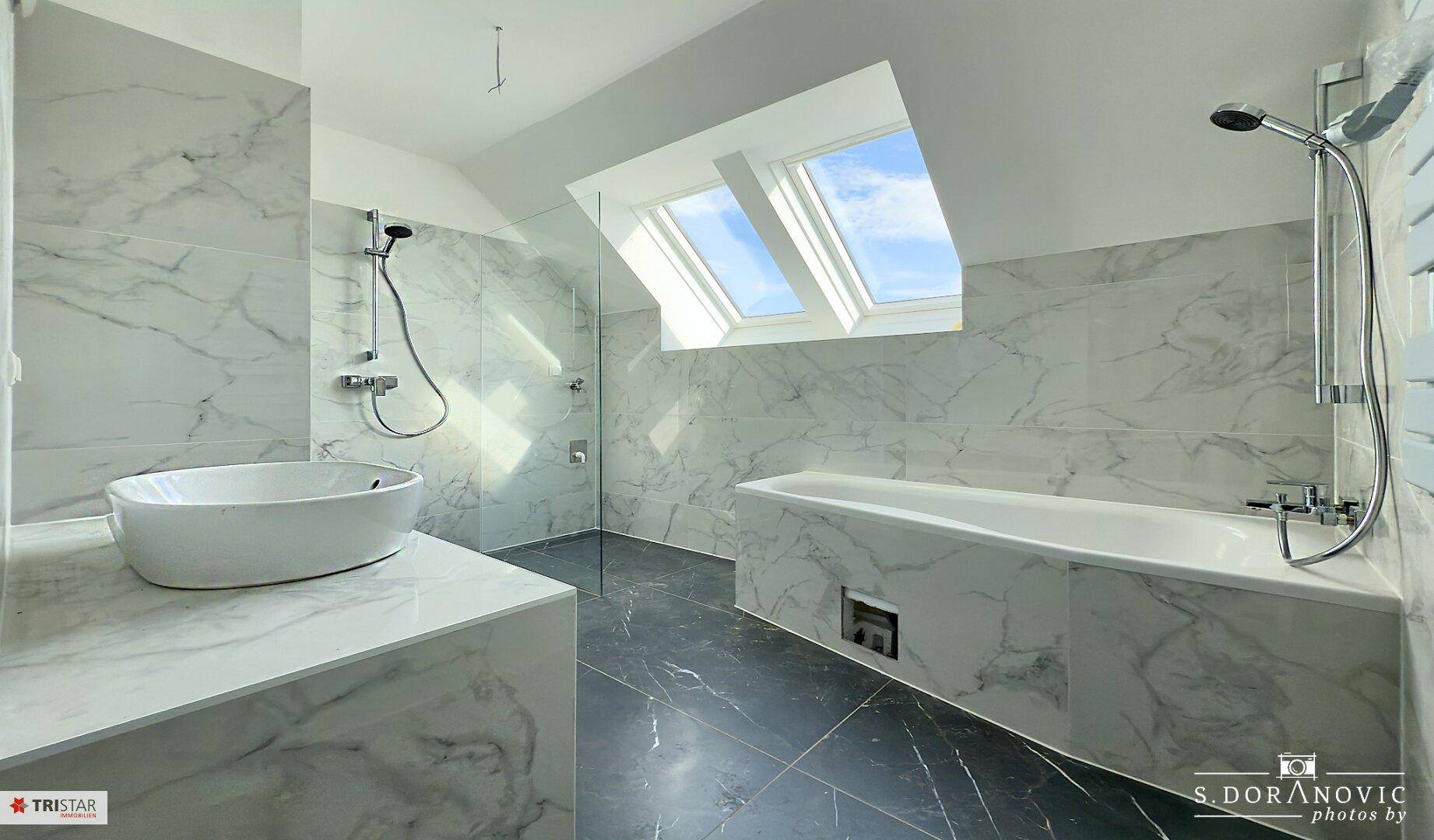 Geräumiges Badezimmer mit eleganten Marmorfliesen, moderner Badewanne und Dusche sowie viel Tageslicht durch das Dachfenster.
