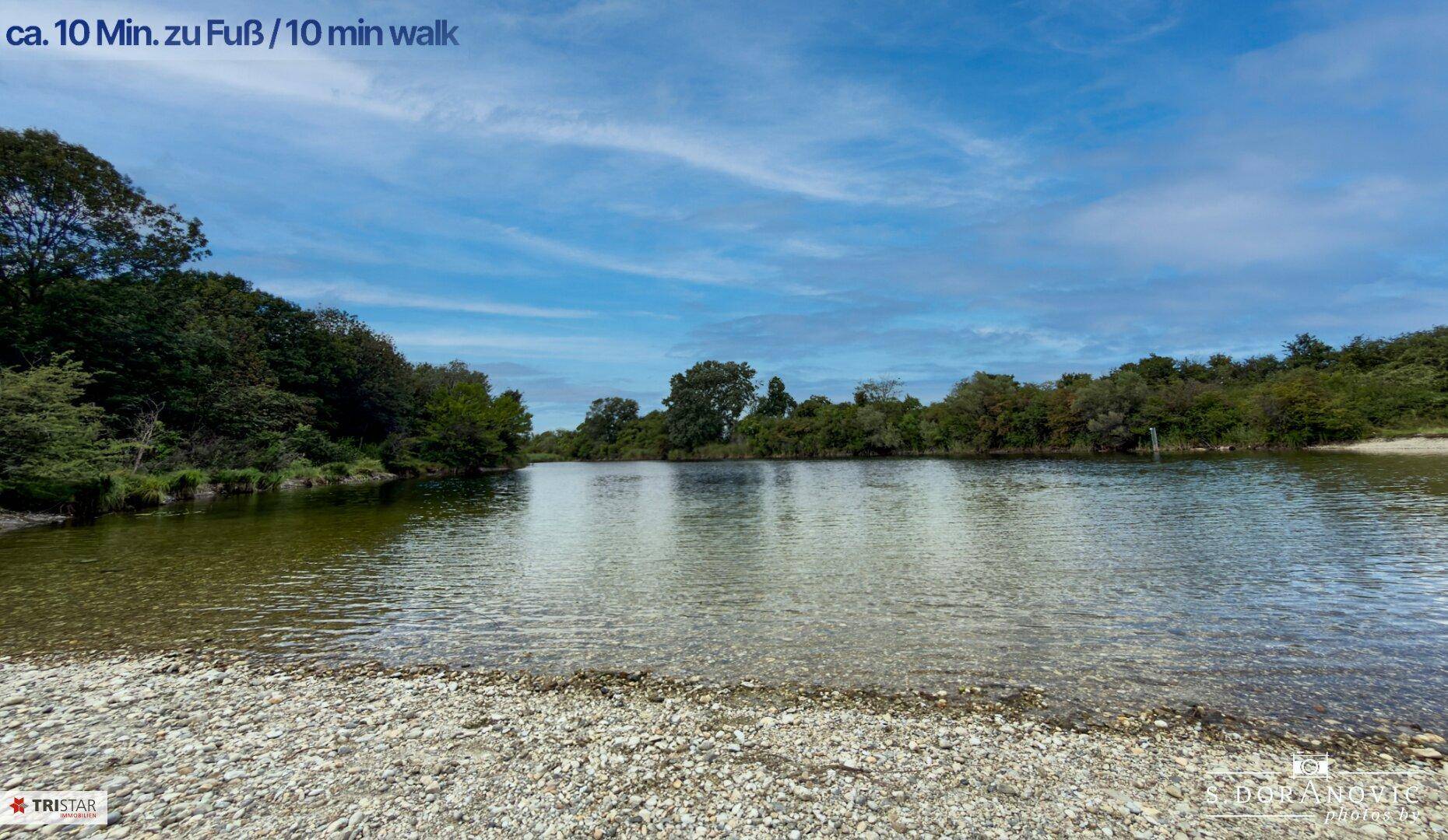 Ruhiger See oder breiter Fluss mit klarem Wasser und bewaldeten Ufern unter blauem Himmel.