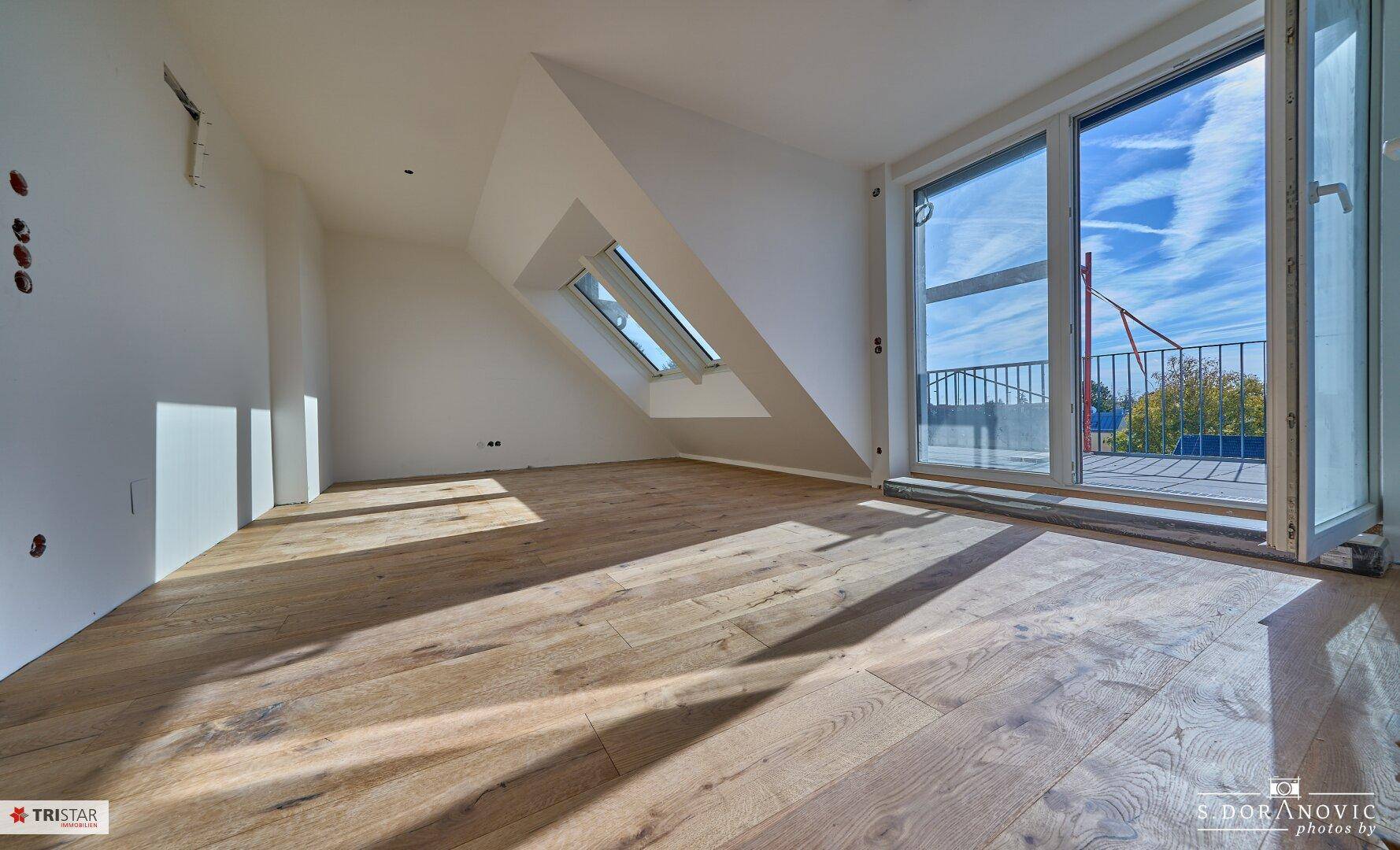 Helles, unmöbliertes Zimmer im Dachgeschoss mit Holzboden und Zugang zu einem Balkon.