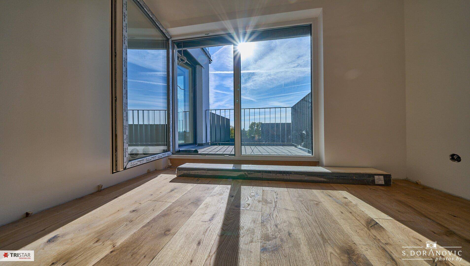 Heller Raum mit Holzboden und großem Fenster, das viel Sonnenlicht hereinlässt.
