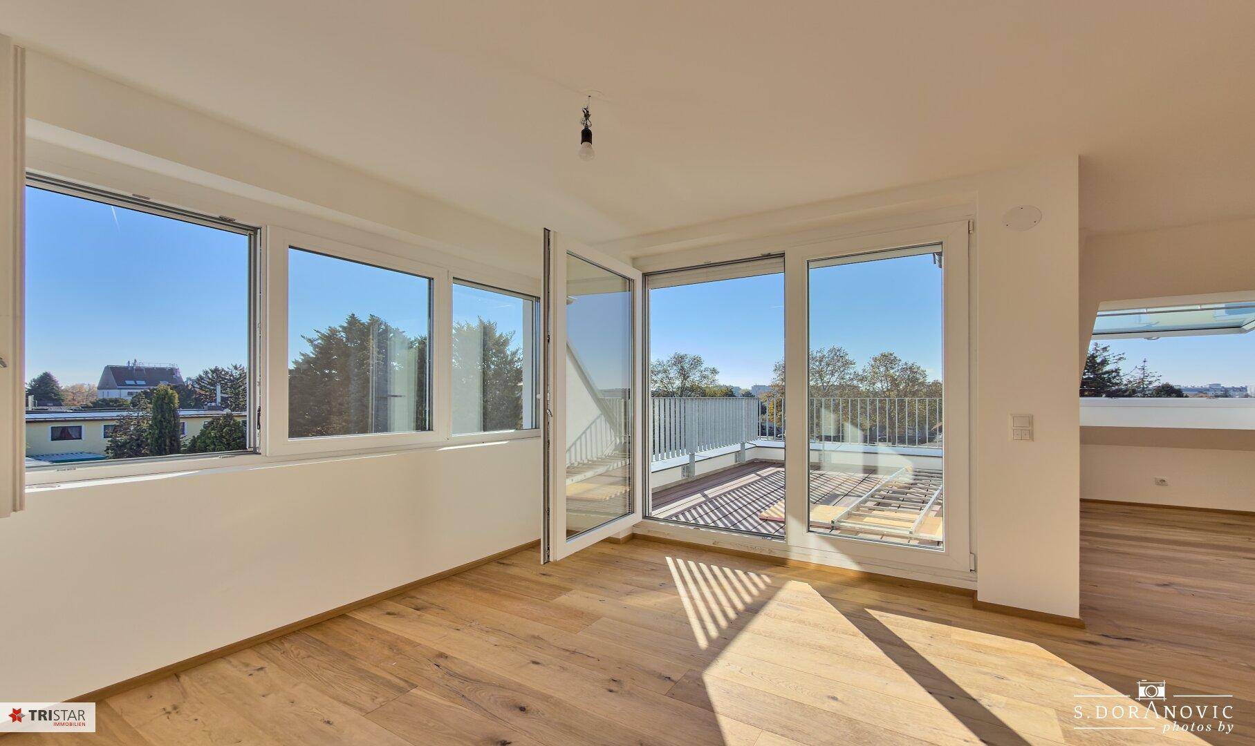 Heller, unmöblierter Wohnbereich mit großen Fenstern, Holzboden und Zugang zur Terrasse.