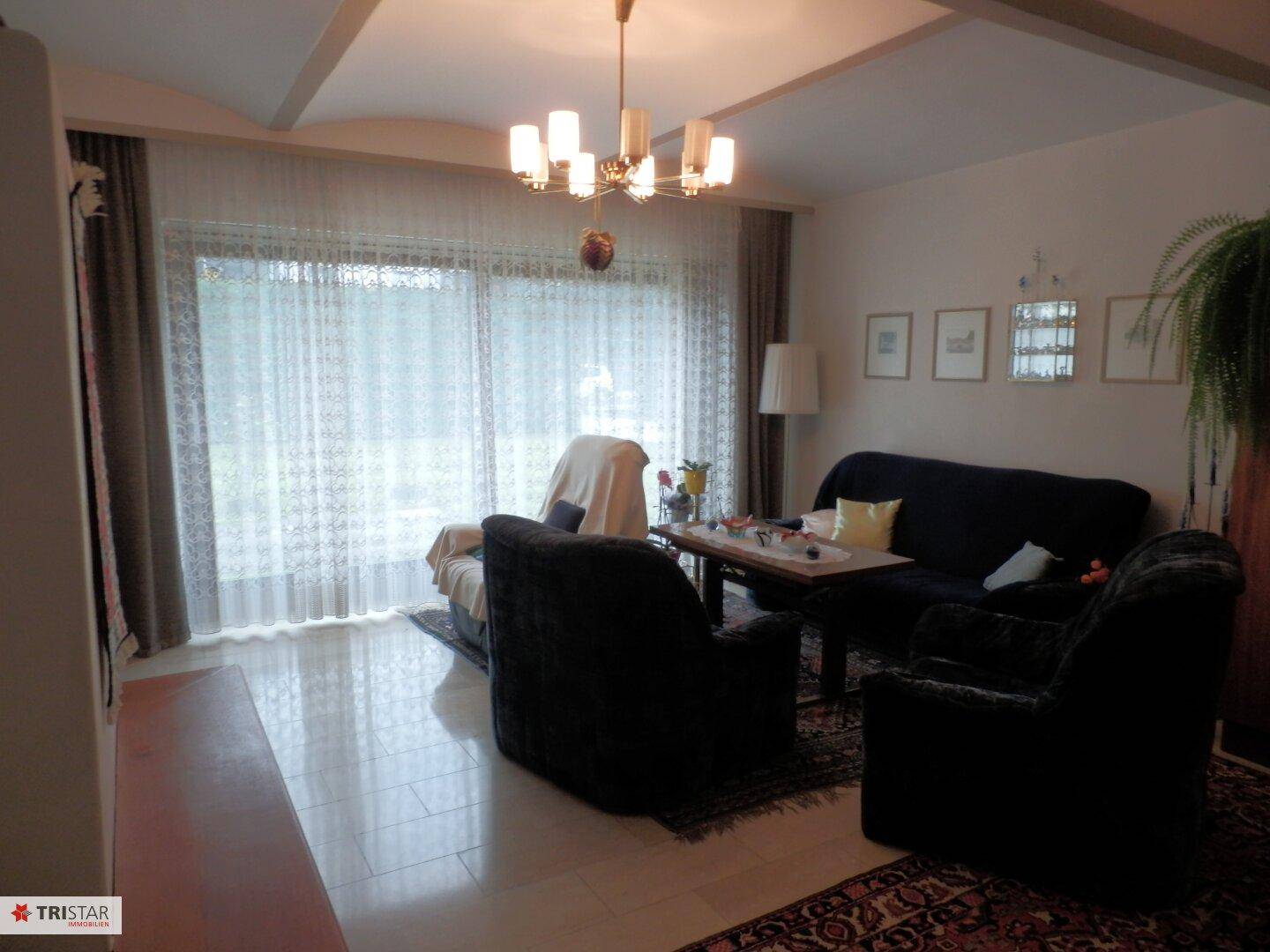 Helles Wohnzimmer mit Fliesenboden, großen Fenstern und Vorhängen.