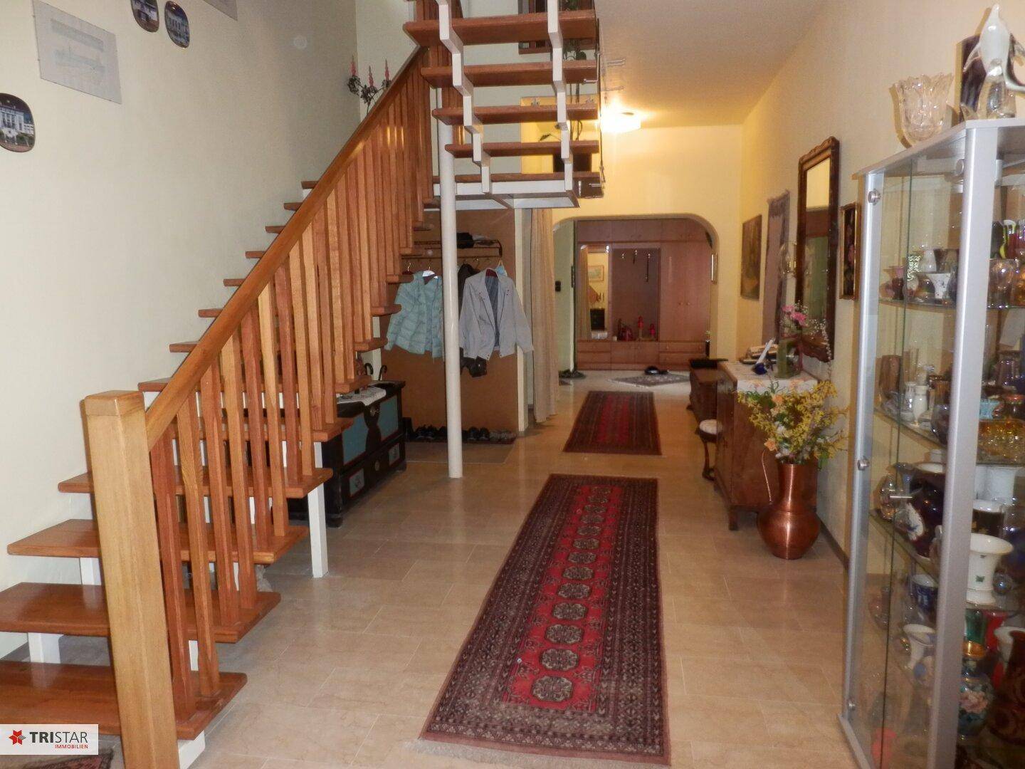 Geräumiger Flur mit Holztreppe, Fliesenboden und Vitrinenschrank.
