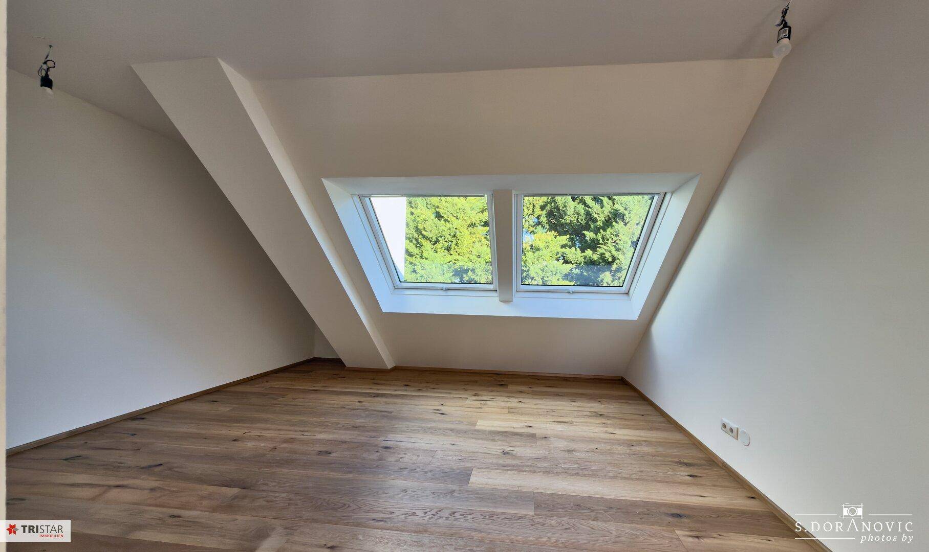 Helles Zimmer mit Holzboden und zwei Dachfenstern, die einen Blick ins Grüne bieten.