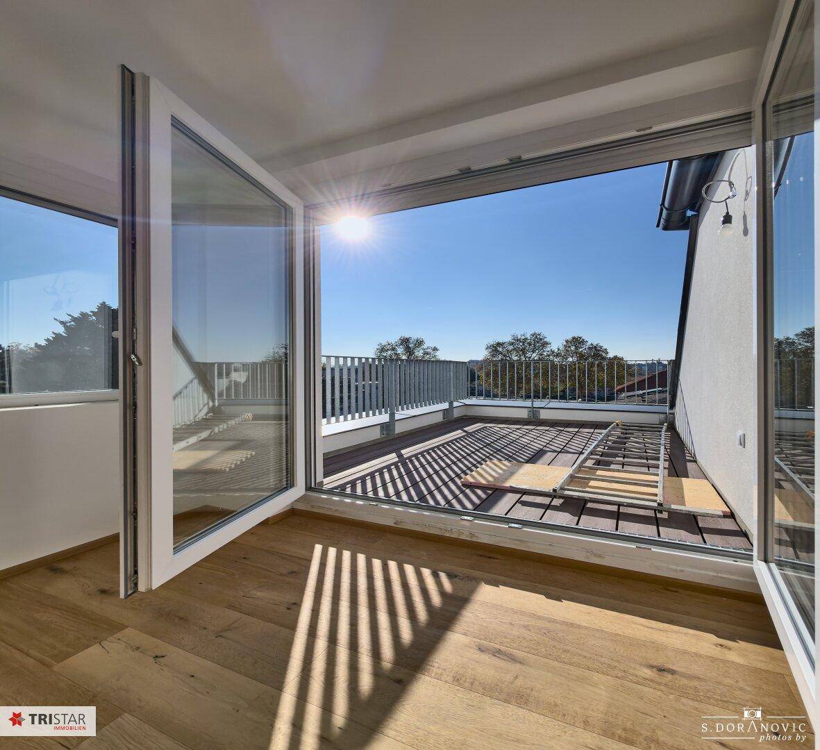Heller Raum mit Holzboden und großen Fenstern, die Zugang zu einer sonnigen Dachterrasse bieten.