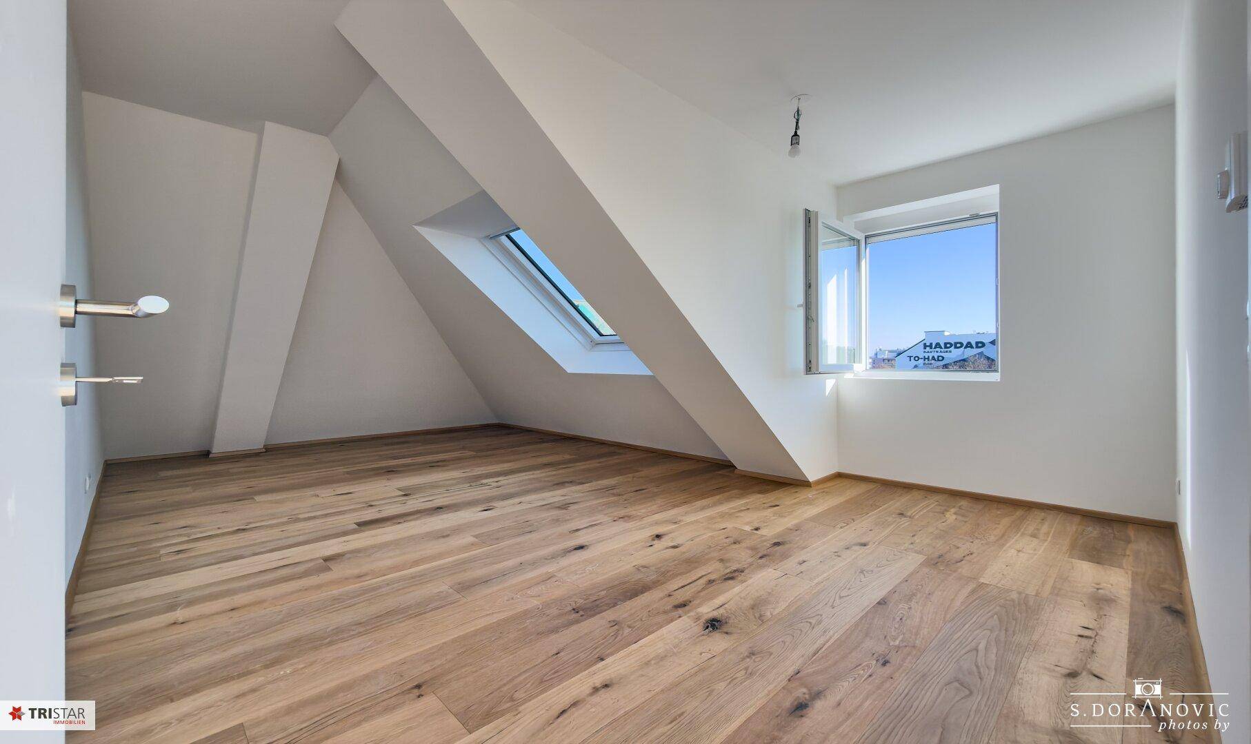 Geräumiges Zimmer mit Holzboden, Dachfenster und einem normalen Fenster mit Ausblick.