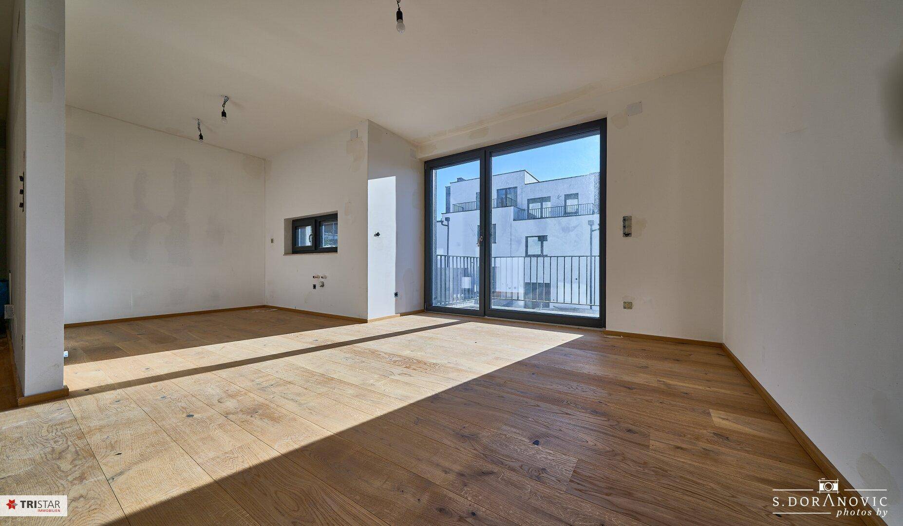 Helles, leeres Zimmer mit Holzboden und großen Schiebetüren, die auf einen Balkon führen.