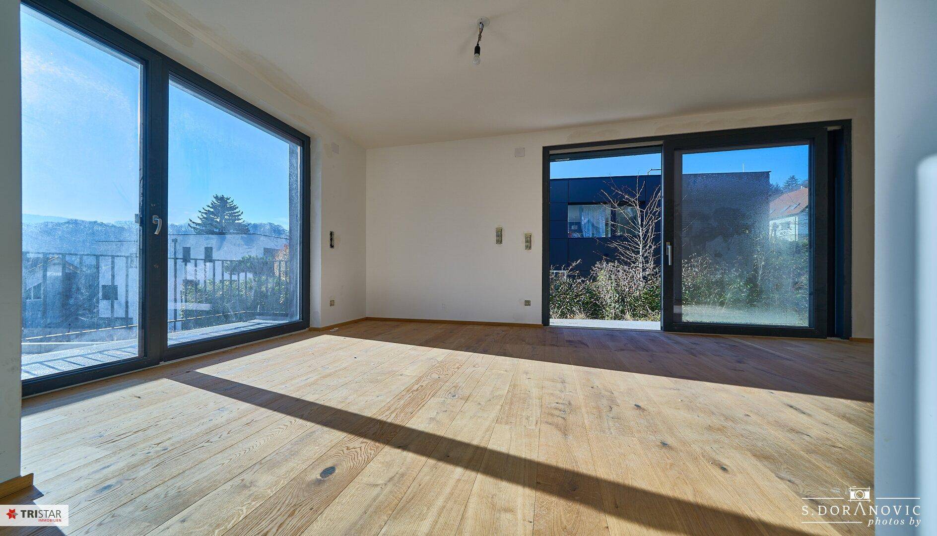 Geräumiges Zimmer mit Holzboden und großen Fenstern, die viel Tageslicht hereinlassen und eine grüne Aussicht bieten.