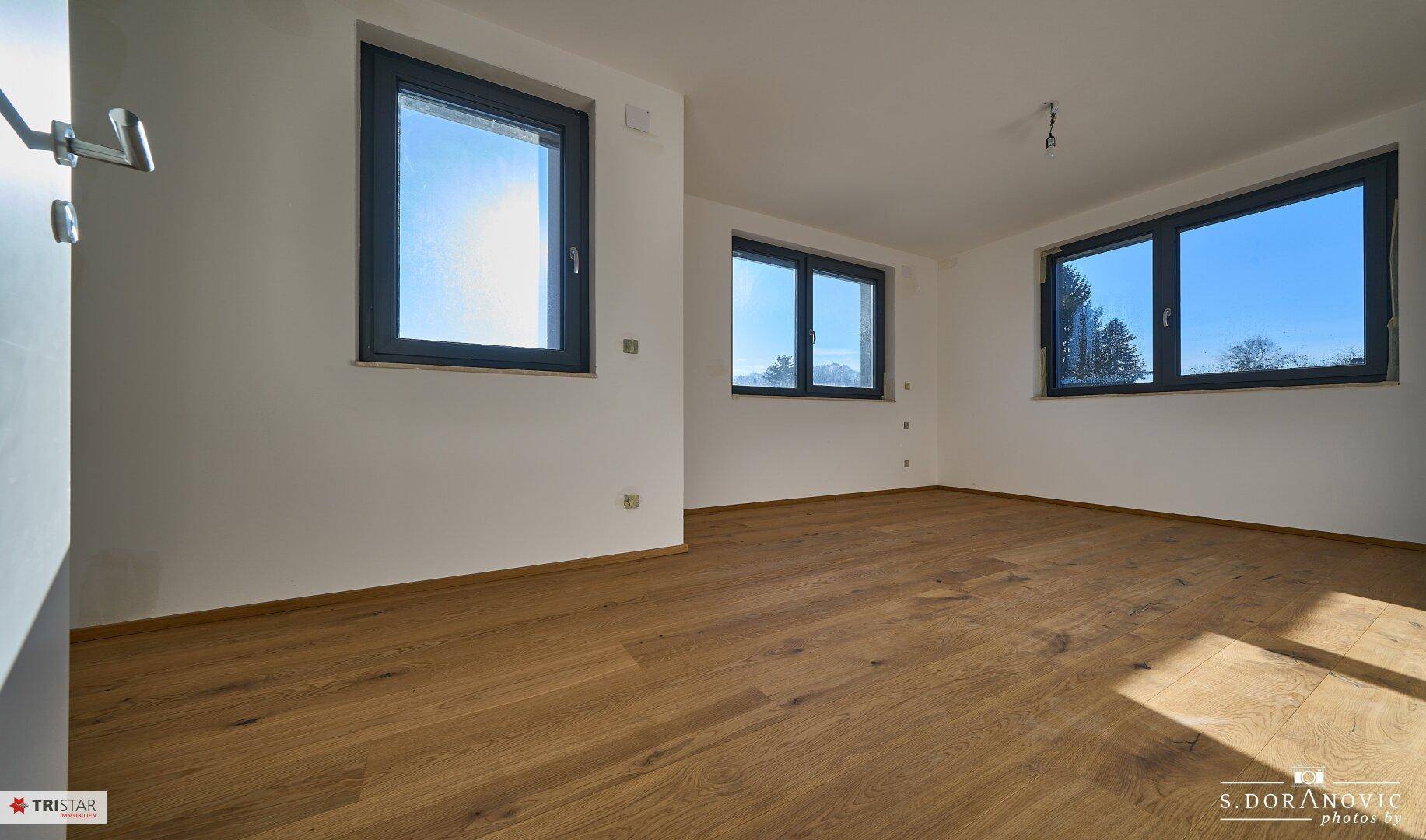 Helles Zimmer mit Holzboden und mehreren Fenstern, die einen weiten Blick in die Umgebung ermöglichen.