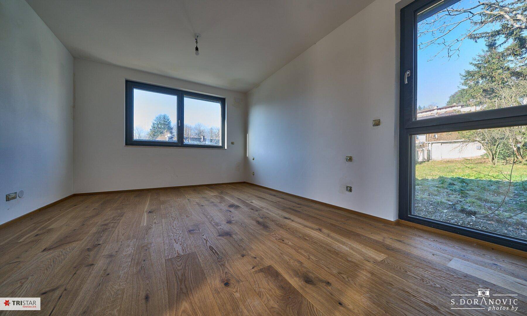 Helles Zimmer mit Holzboden und Fenstern, die einen Blick auf den Garten und die umliegende Natur bieten.