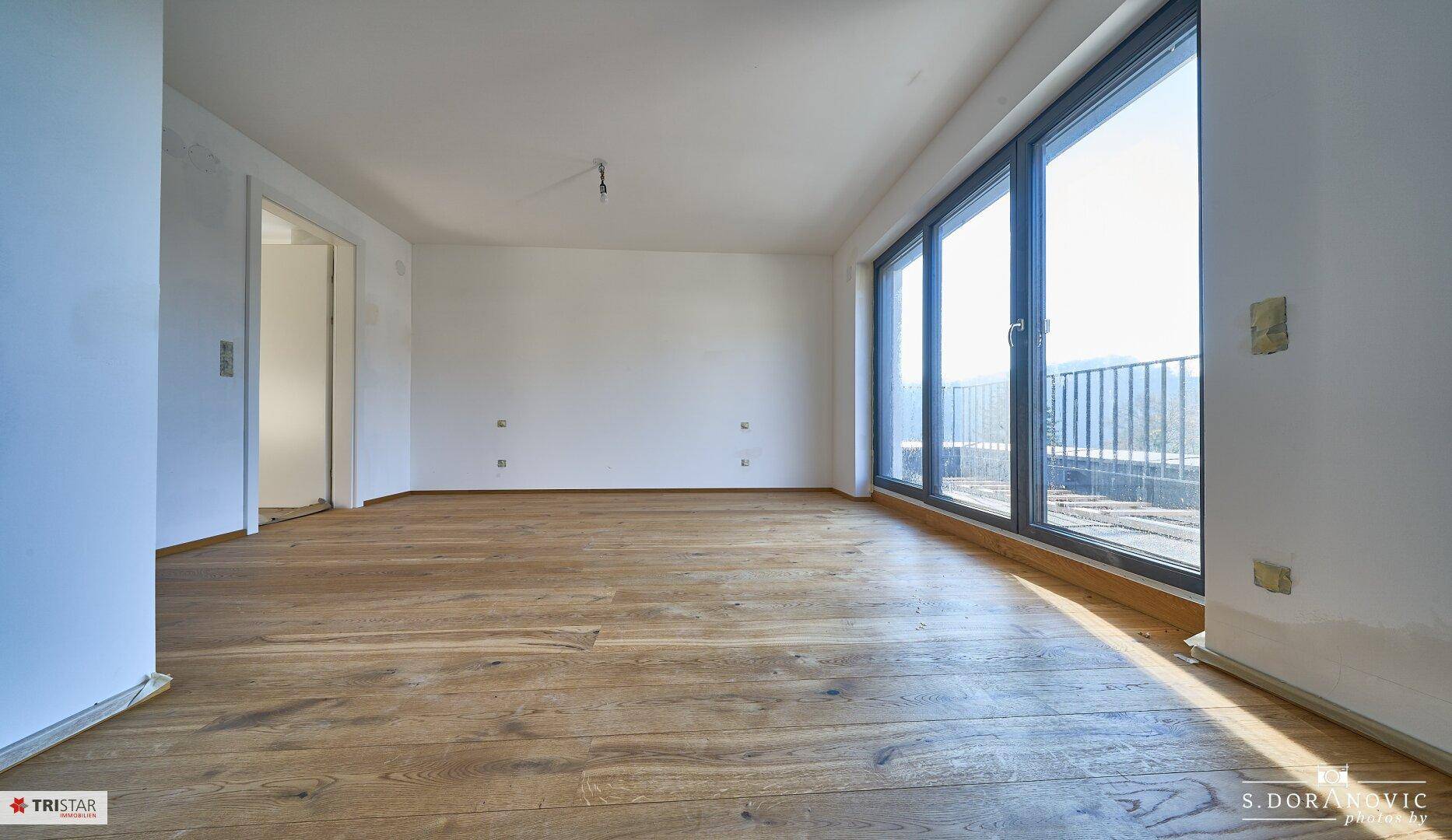 Helles Zimmer mit Holzboden und einer Fensterfront, die einen weiten Blick in die Landschaft ermöglicht.