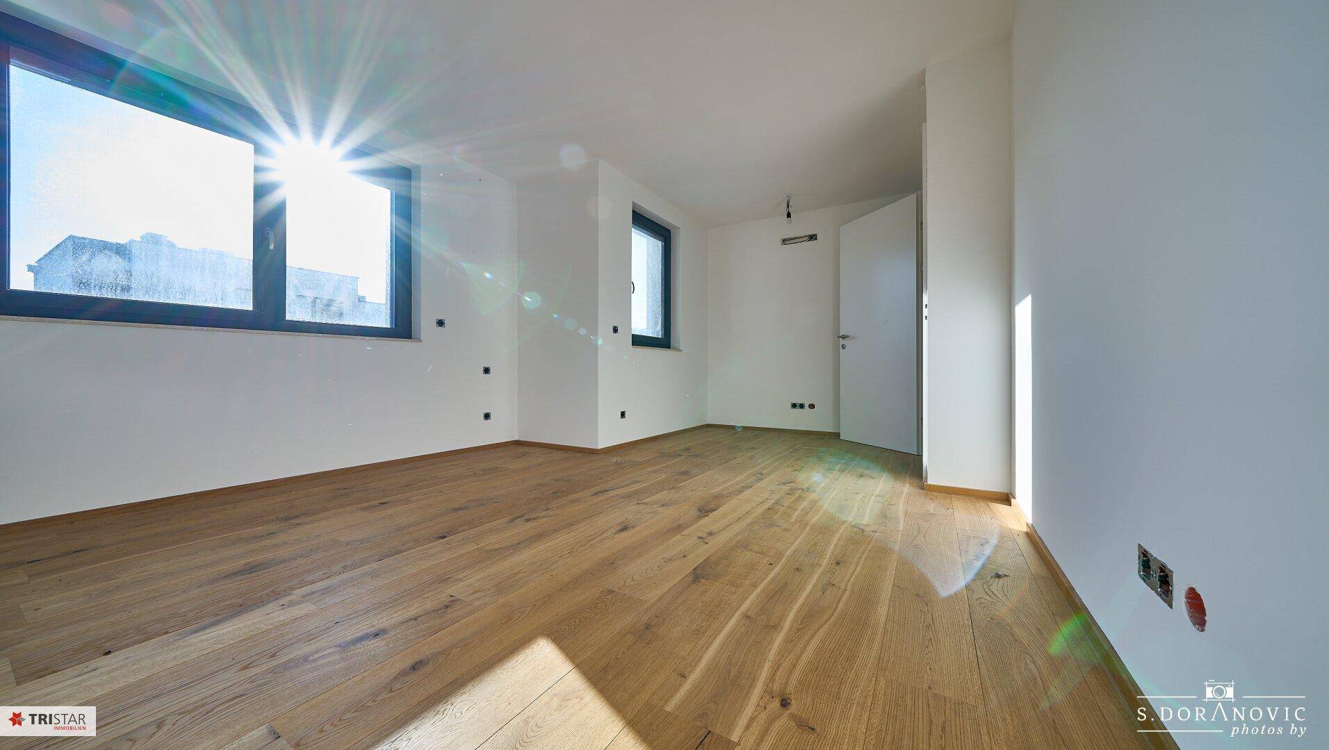 Lichtdurchflutetes Zimmer mit Holzboden und großen Fenstern, die den Raum erhellen.