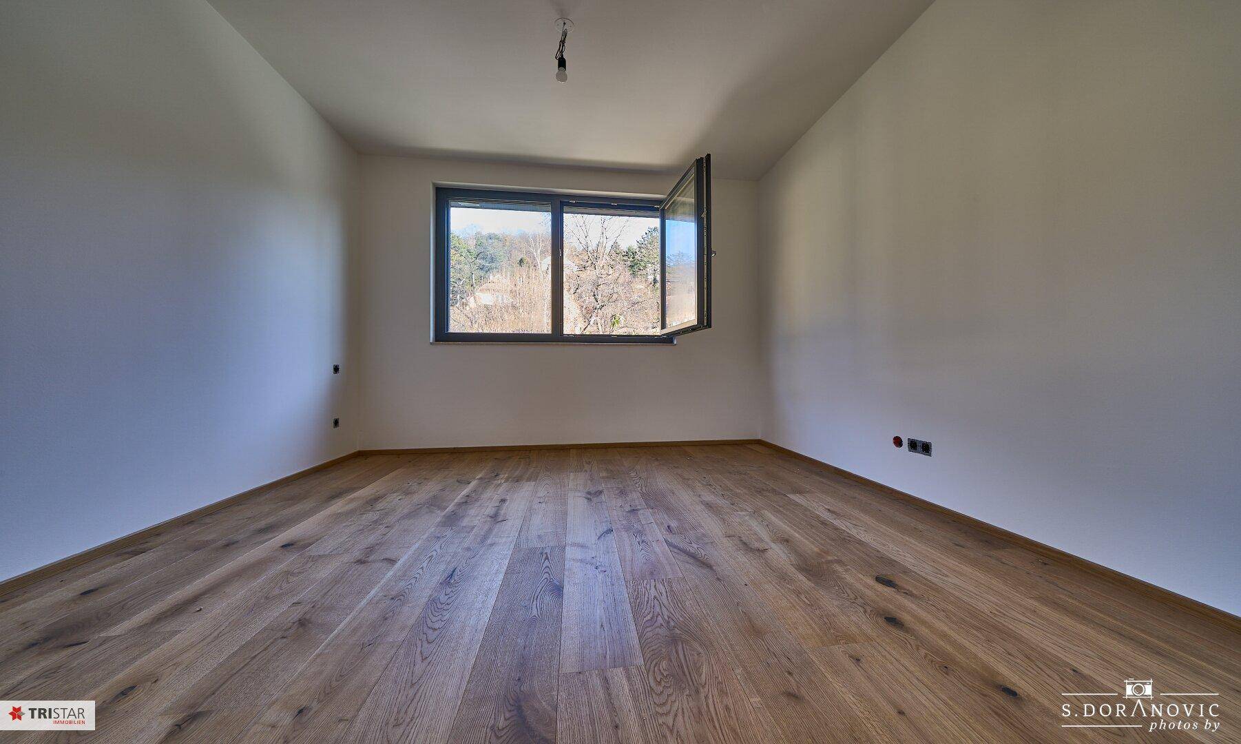 Helles Schlafzimmer mit Holzboden und einem geöffneten Fenster, das einen Blick ins Grüne bietet.