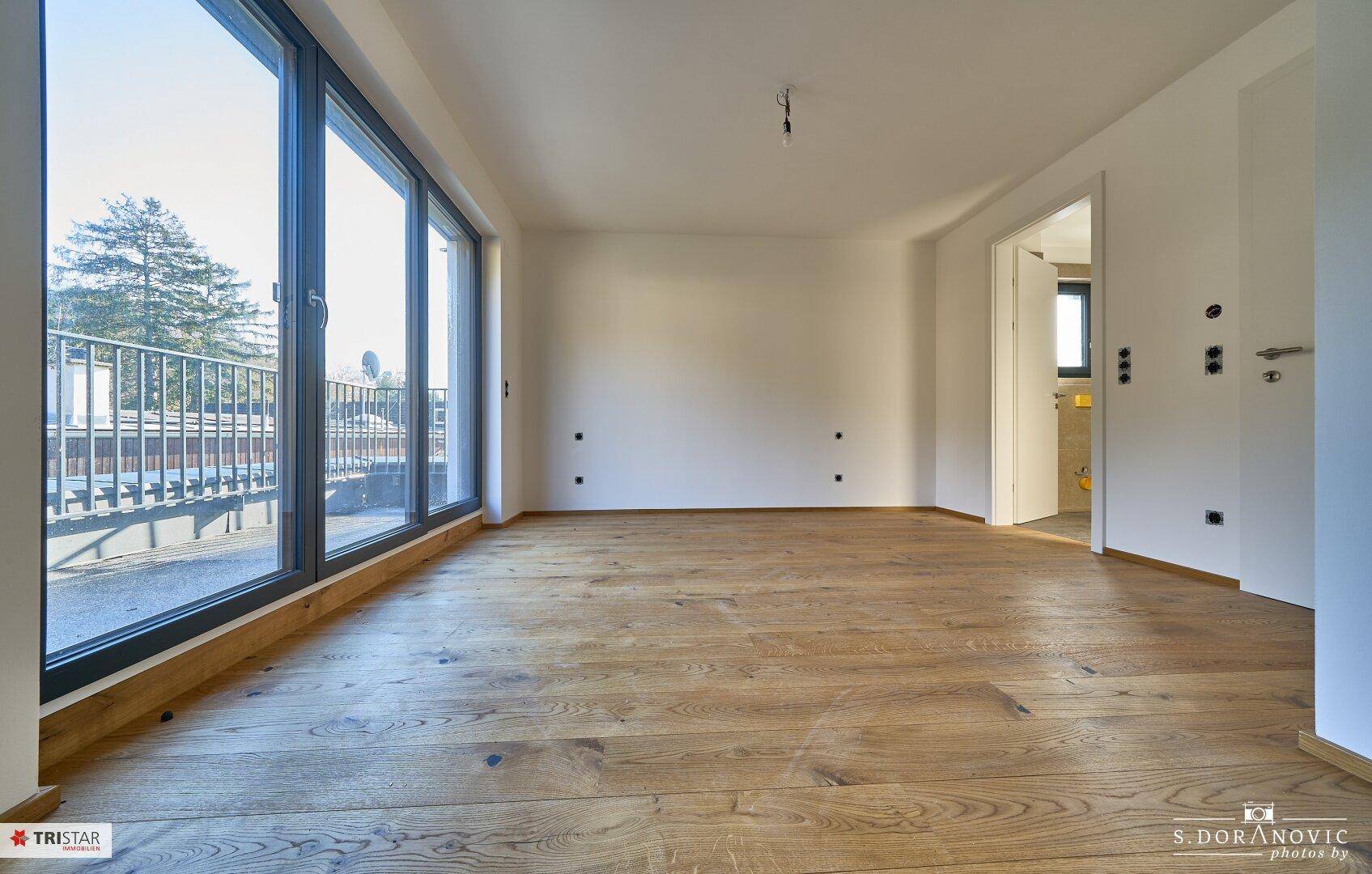 Heller Wohnraum mit Holzboden und bodentiefen Fenstern, die Zugang zu einem Balkon bieten.