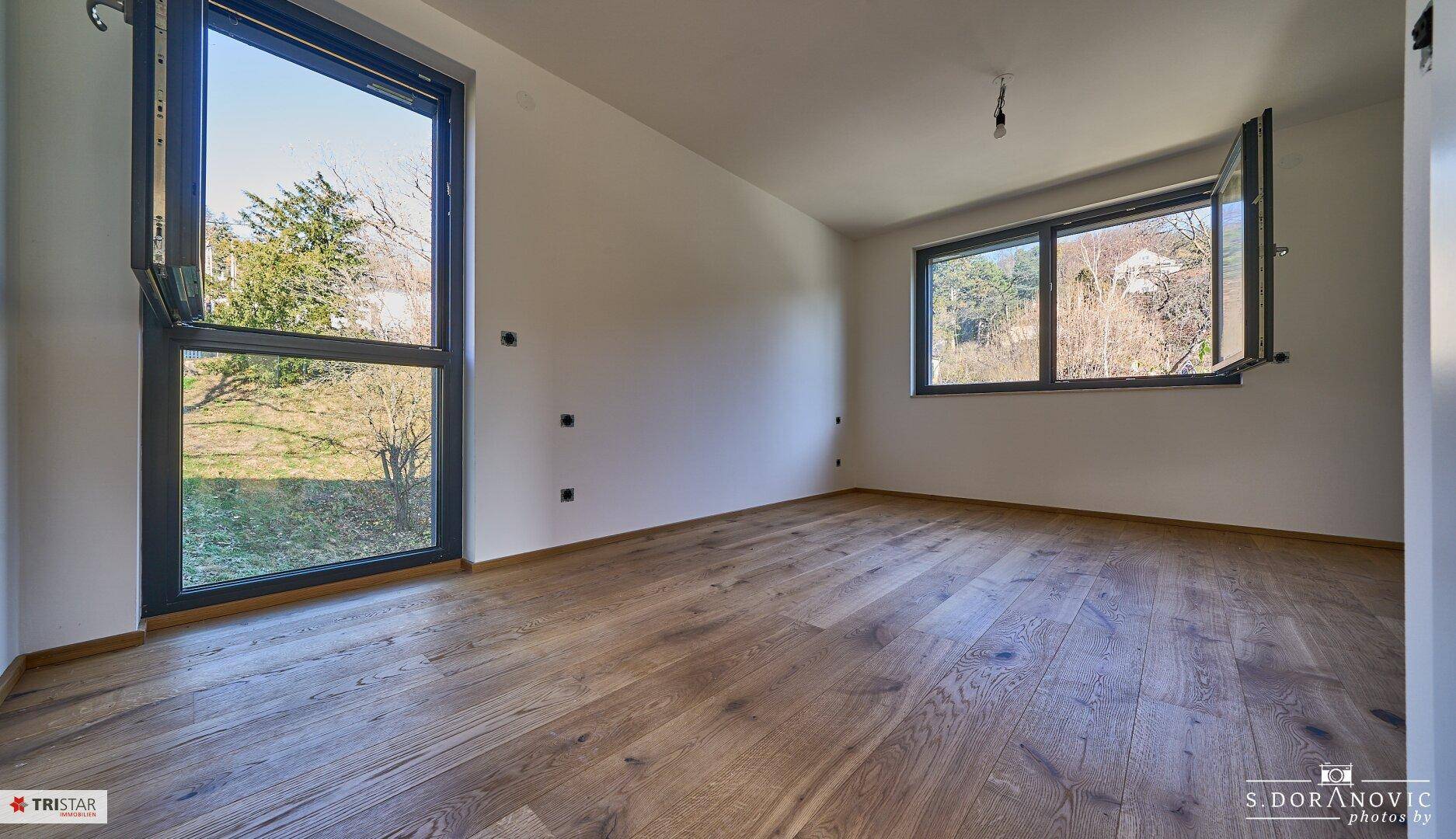 Helles Schlafzimmer mit Holzboden und großen Fenstern mit Blick ins Grüne.