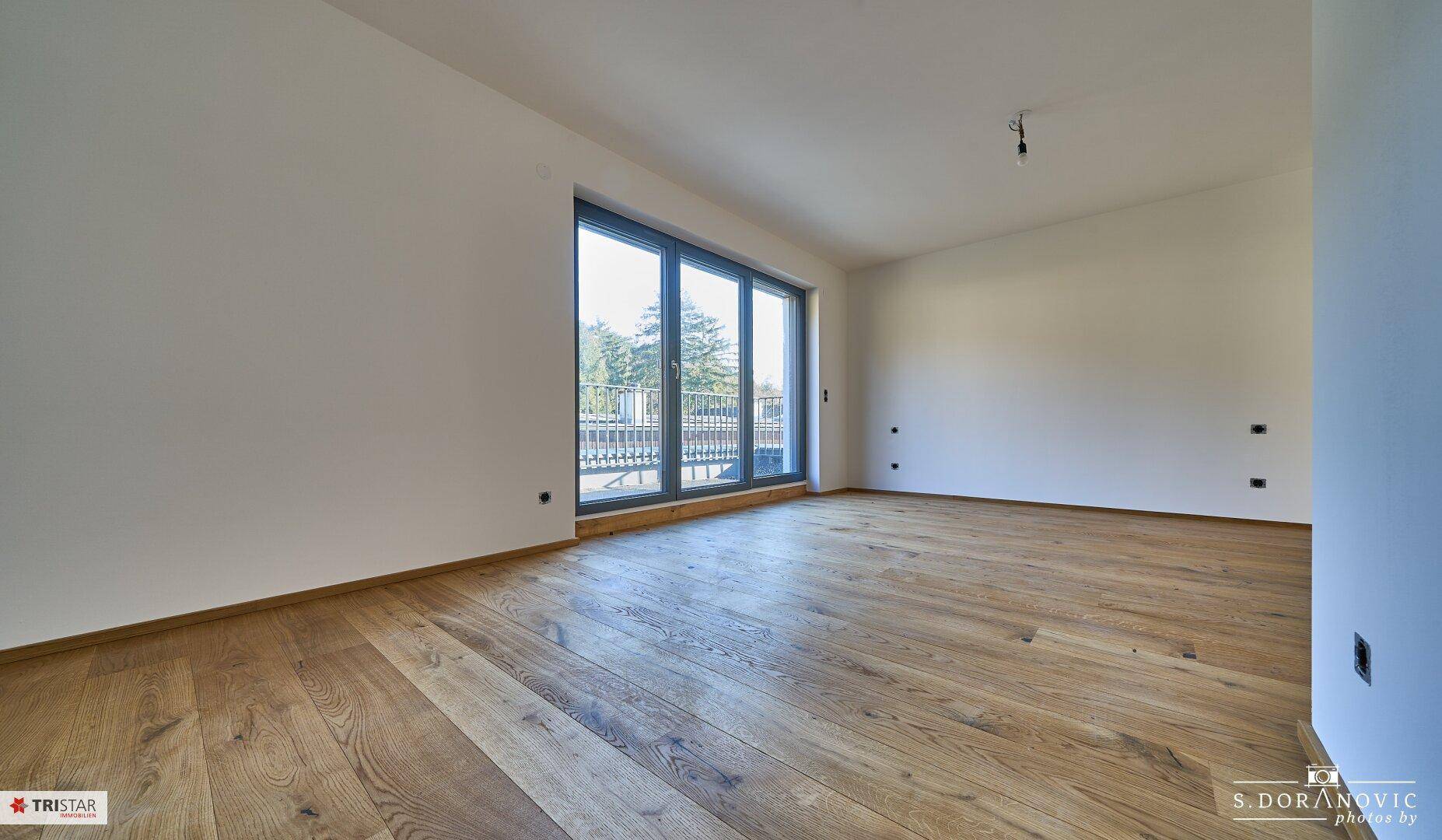 Geräumiger, heller Wohnbereich mit Holzboden und bodentiefen Fenstern, bereit für die Einrichtung.