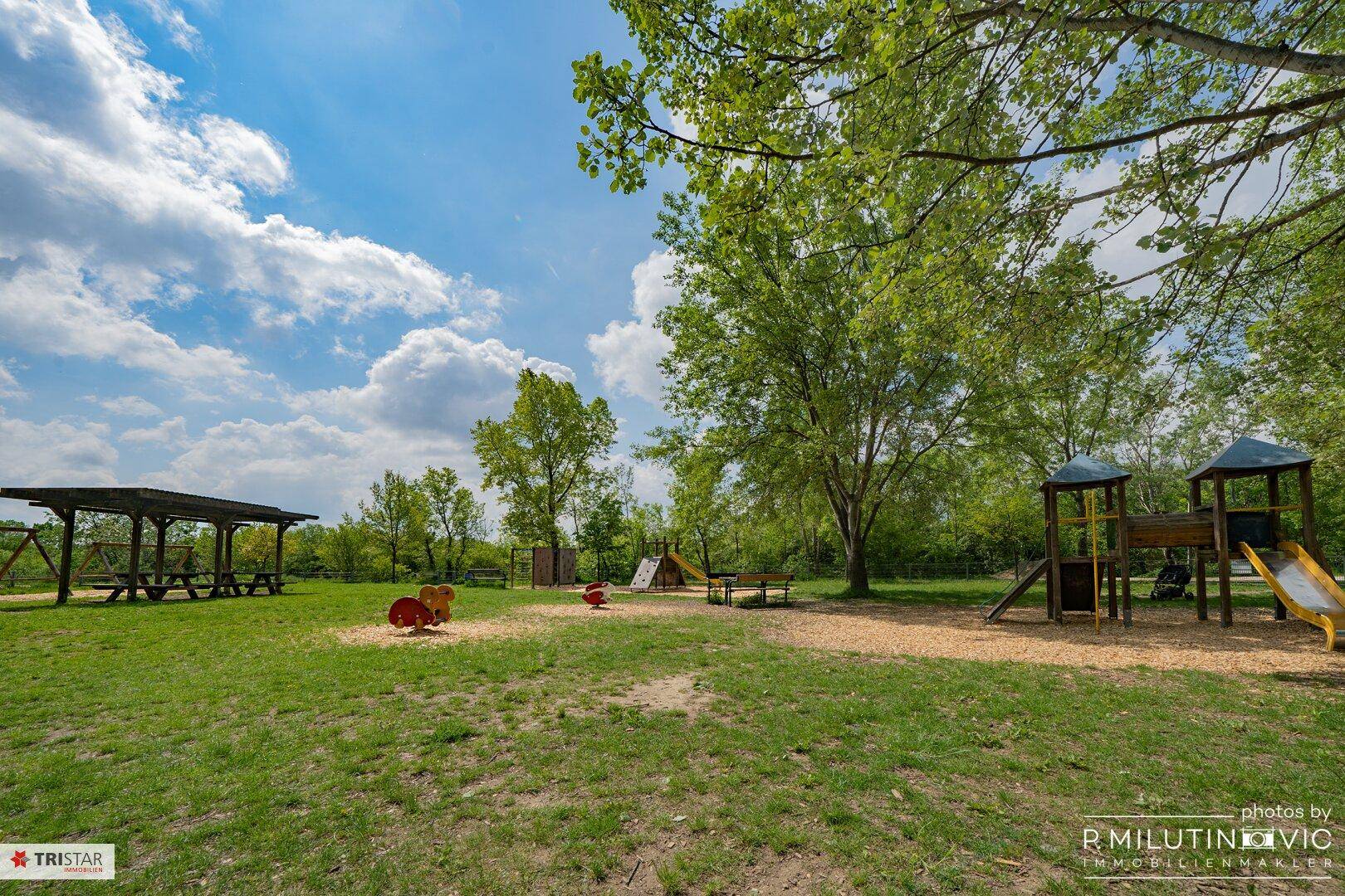 Großer Spielplatz mit Schaukeln, Rutschen und einem überdachten Picknickbereich im Grünen.