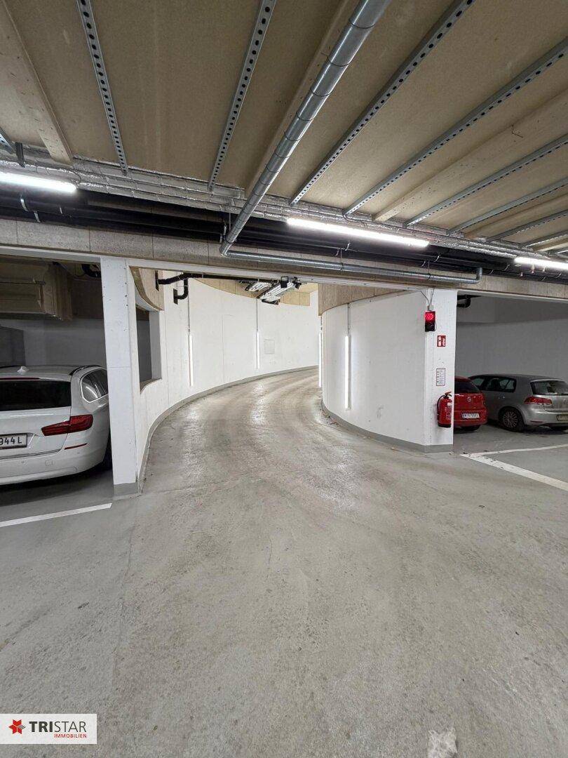 Kurvige Rampe in einer Tiefgarage mit geparkten Fahrzeugen und klarer Beschilderung.