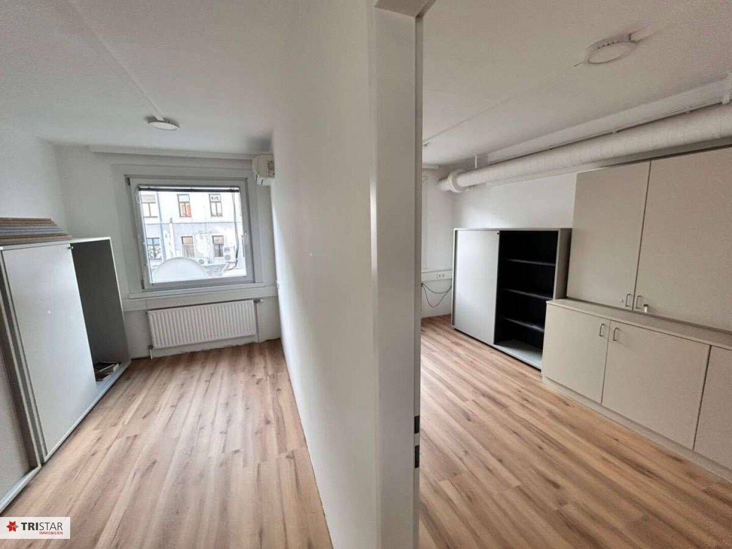 Büroraum mit Laminatboden und Einbauschränken, geteilt durch eine Säule, mit Fensterblick.