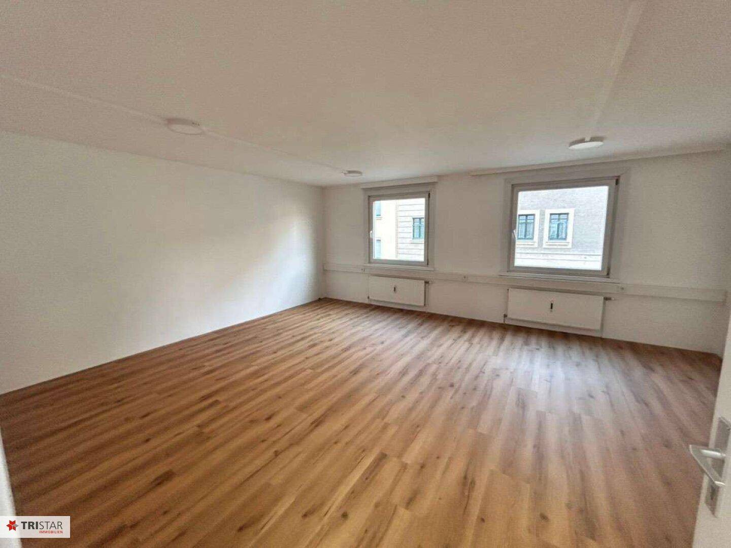 Geräumiger Büroraum mit Laminatboden und zwei Fenstern, ideal für verschiedene Arbeitsbereiche.
