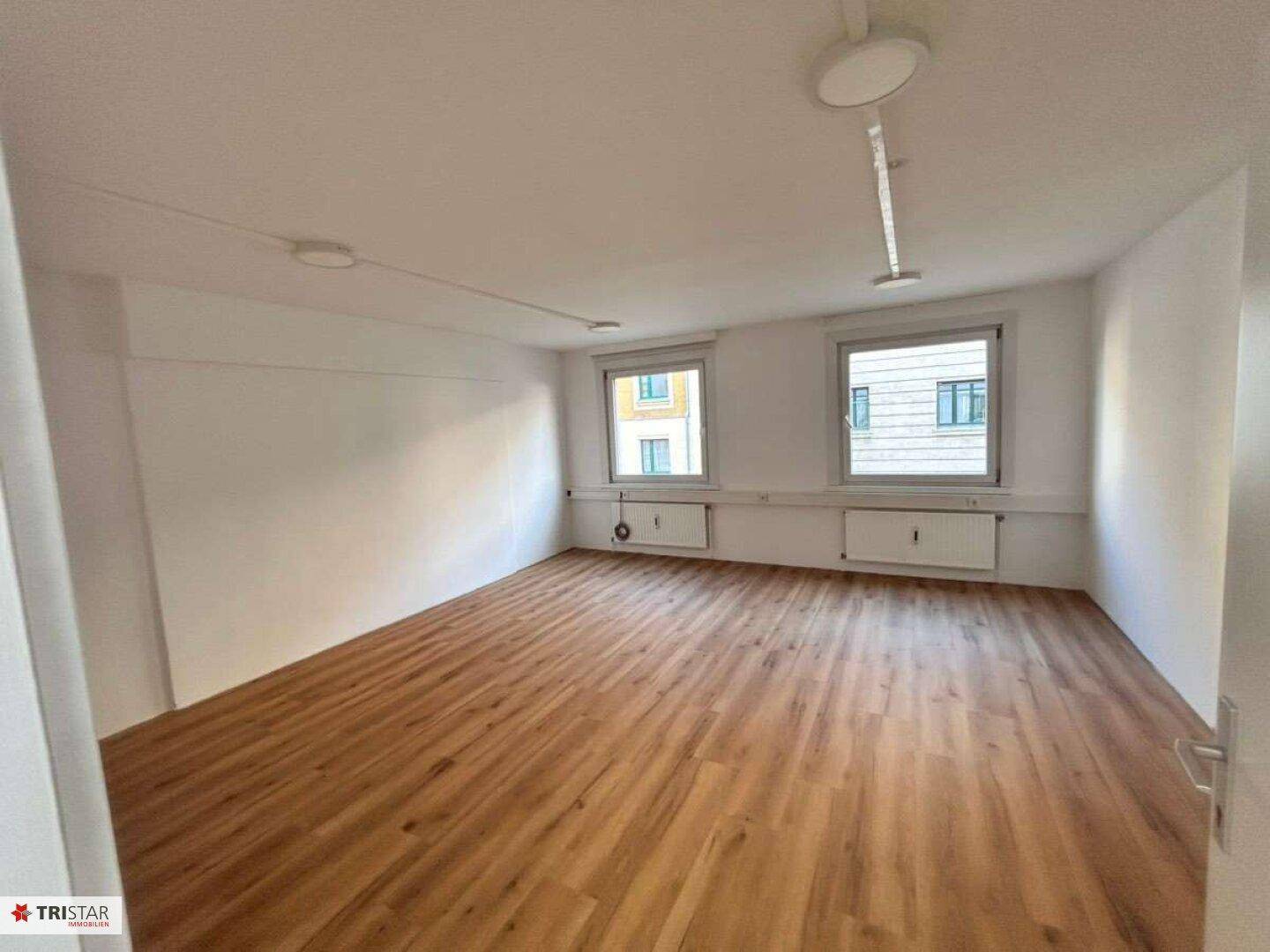 Heller Büroraum mit Laminatboden und zwei Fenstern, die eine angenehme Arbeitsatmosphäre schaffen.