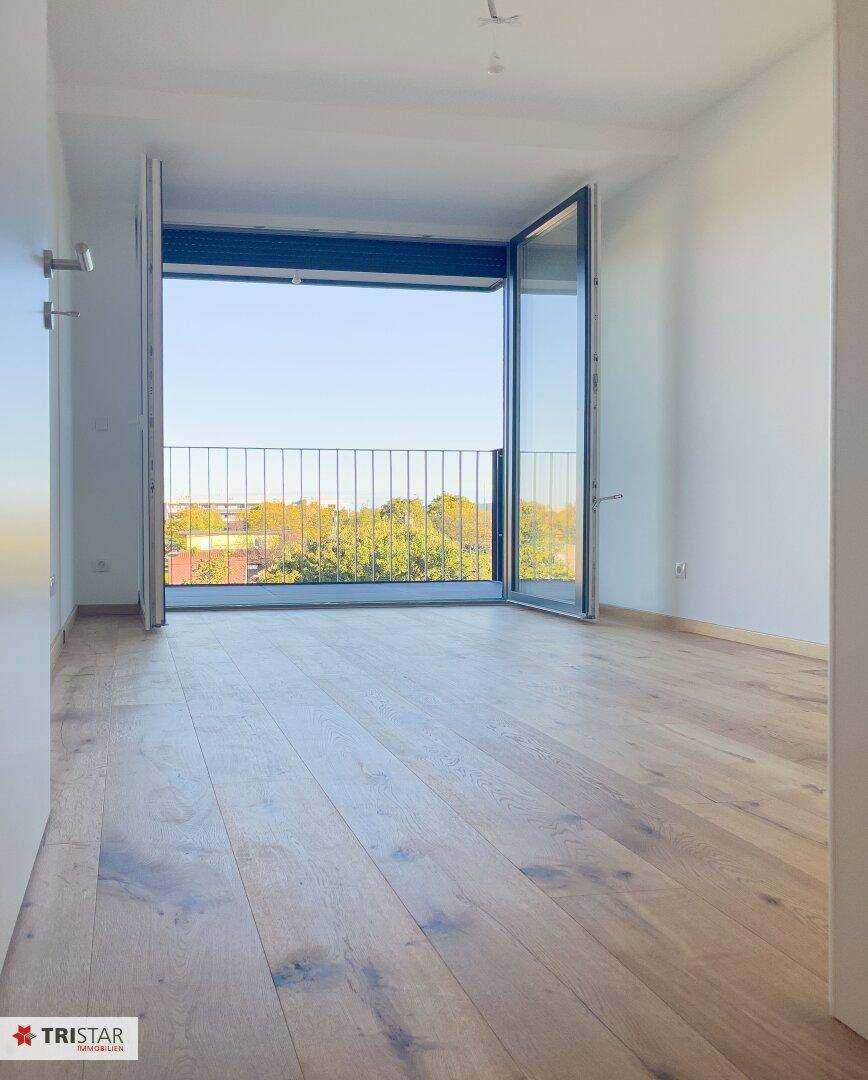 Leeres Schlafzimmer mit Holzboden und breiten Fenstern, die Zugang zum Balkon bieten.
