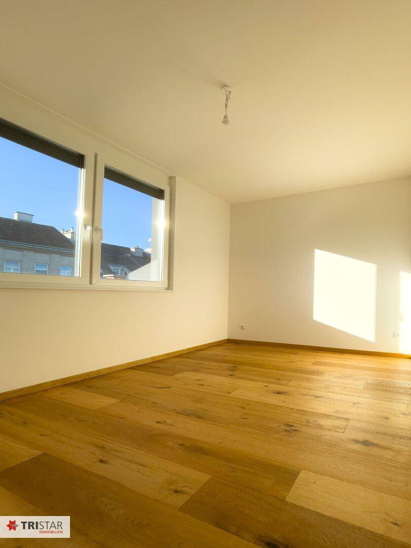 Leeres Schlafzimmer mit Holzboden und zwei Fenstern, die viel Sonnenlicht hereinlassen.