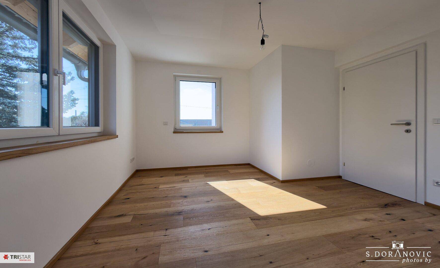 Heller Raum mit Holzboden und zwei Fenstern, eines davon mit Blick ins Grüne.