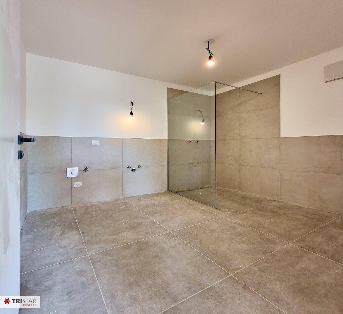 Modernes Badezimmer im Rohbau mit Fliesenboden und vorbereiteter Duschzone.