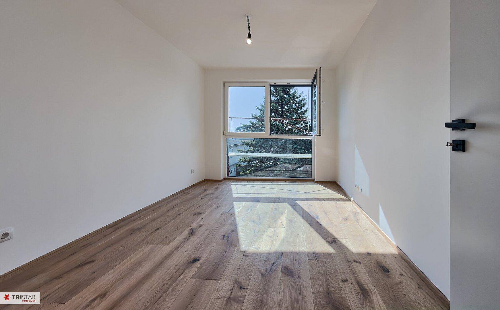 Helles Schlafzimmer mit Holzboden und großem Fenster, das viel Tageslicht hereinlässt.