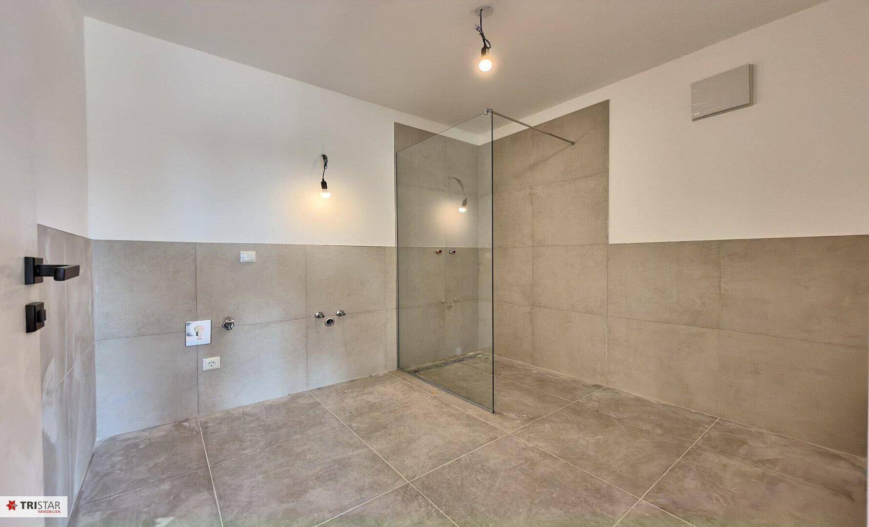 Unfertiges Badezimmer mit modernen Fliesen, Glasduschwand und vorbereiteten Anschlüssen.