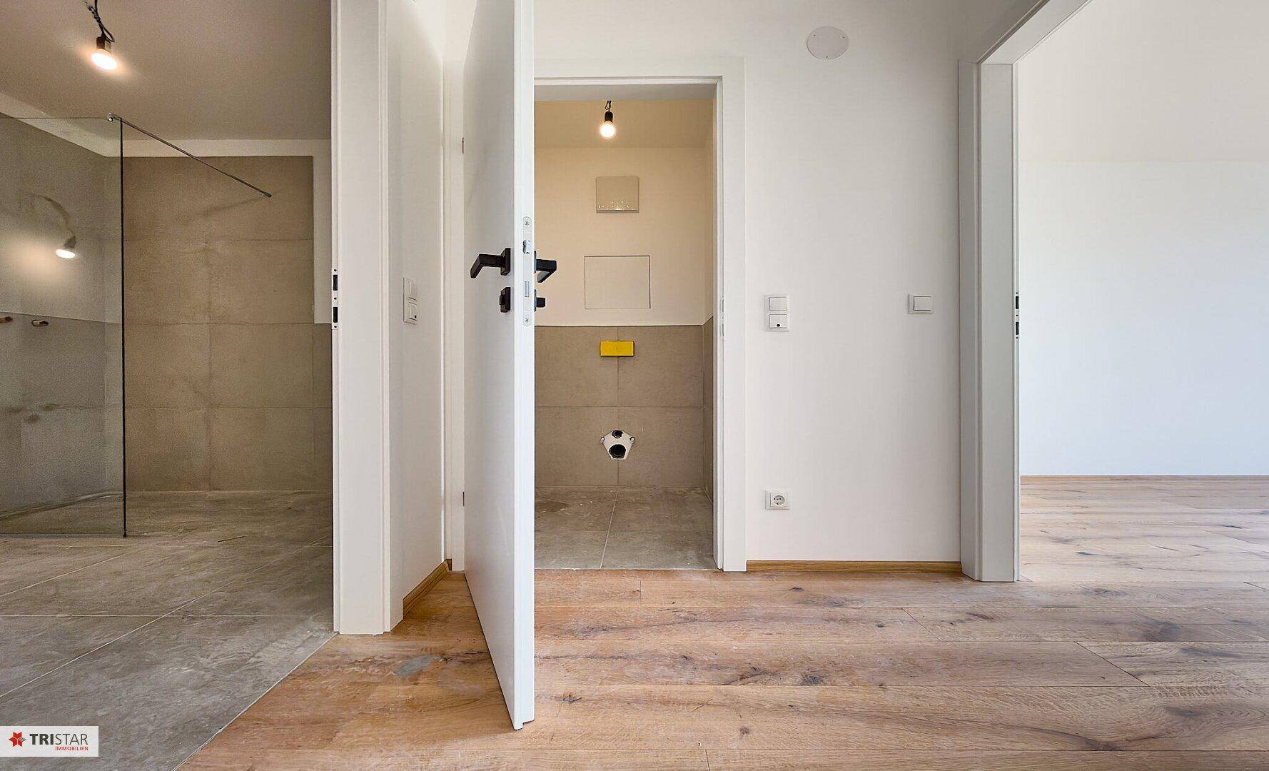 Blick von einem Raum mit Holzfußboden in ein unfertiges WC mit Fliesen und Anschlüssen.