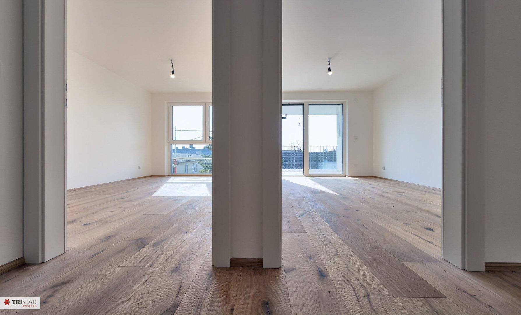 Zwei helle, unmöblierte Räume mit durchgehendem Holzplankenboden und großen Fensterfronten.