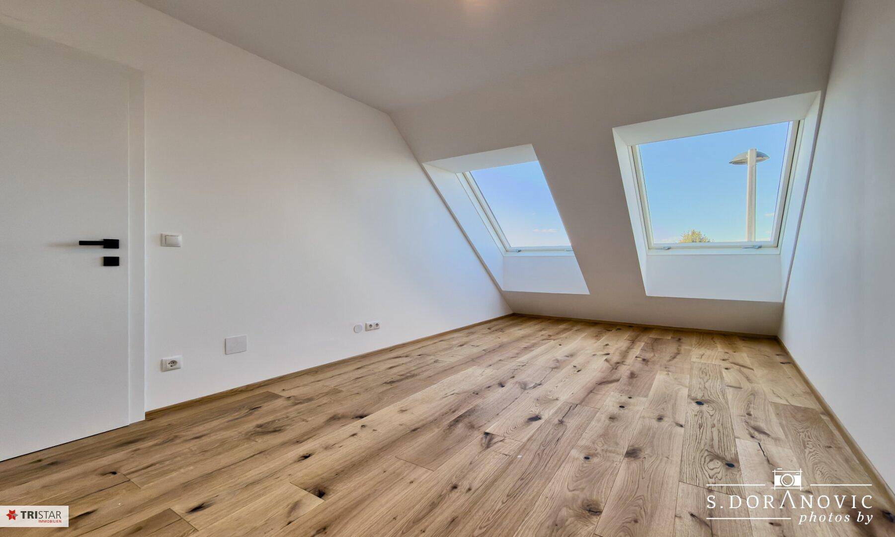 Helles Zimmer mit Holzboden und zwei Dachfenstern in der Dachschräge.
