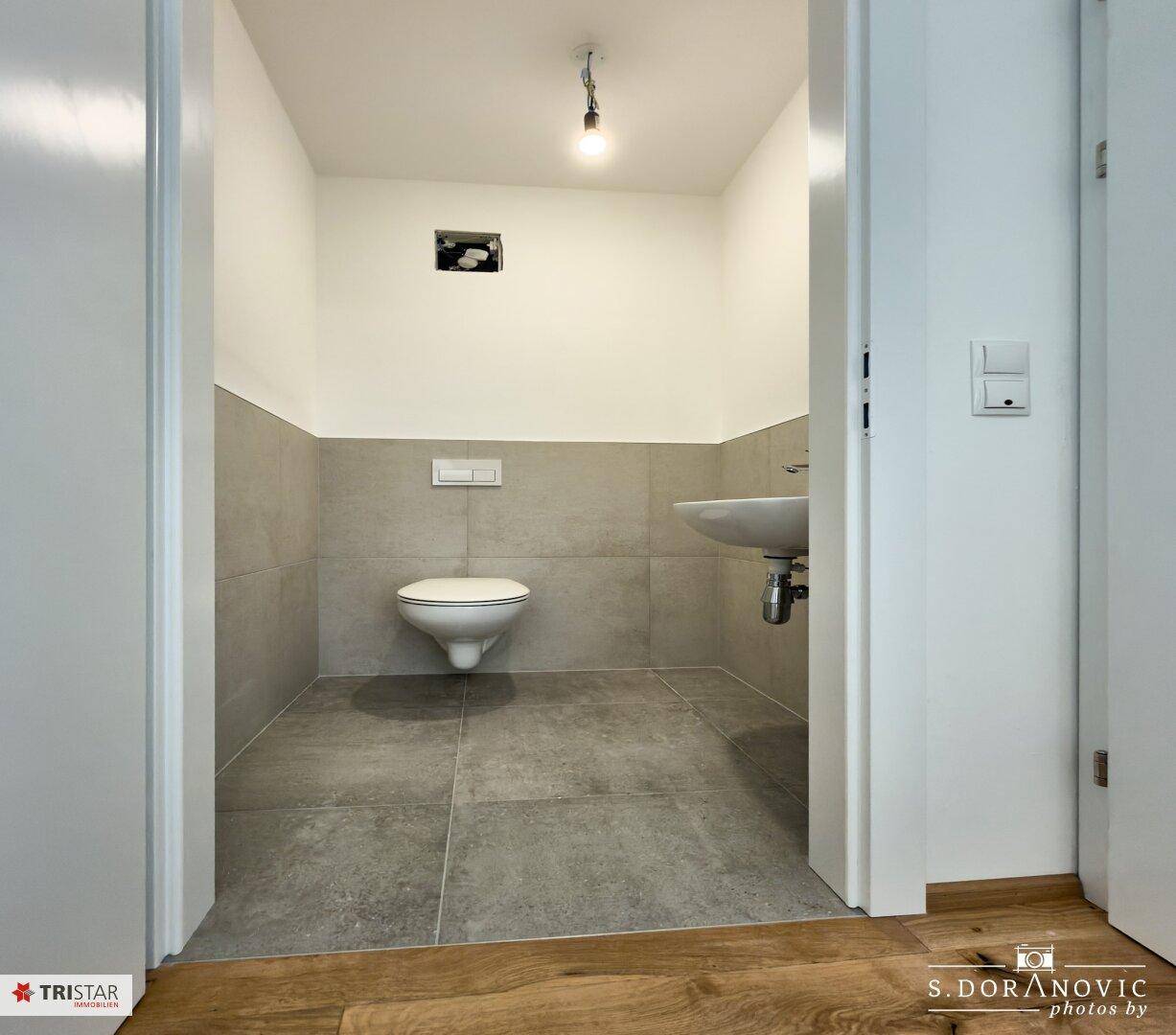 Kleines Gäste-WC mit grauen Fliesen, wandhängender Toilette und Einzelwaschbecken.