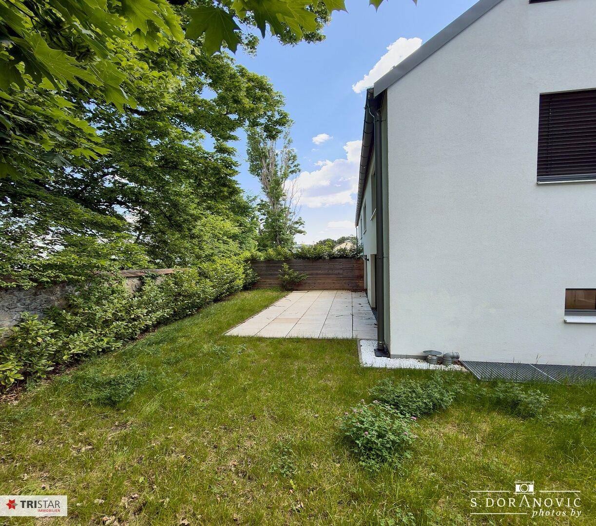 Seitlicher Gartenbereich mit kleiner Terrasse und Rasenfläche, umgeben von Hecken und Bäumen.