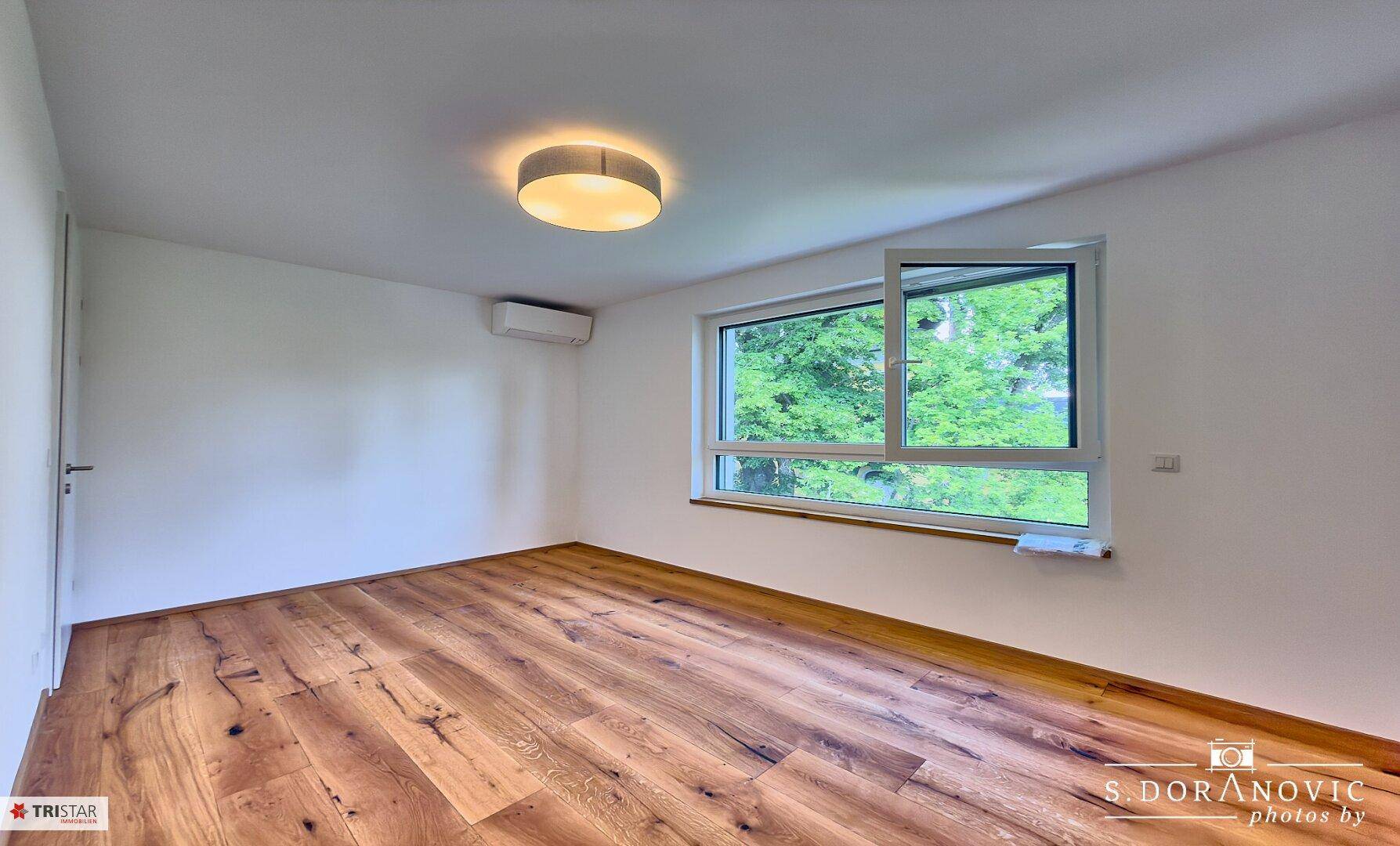 Helles Schlafzimmer mit Holzboden, weißen Wänden und großem Fenster mit Grünblick.