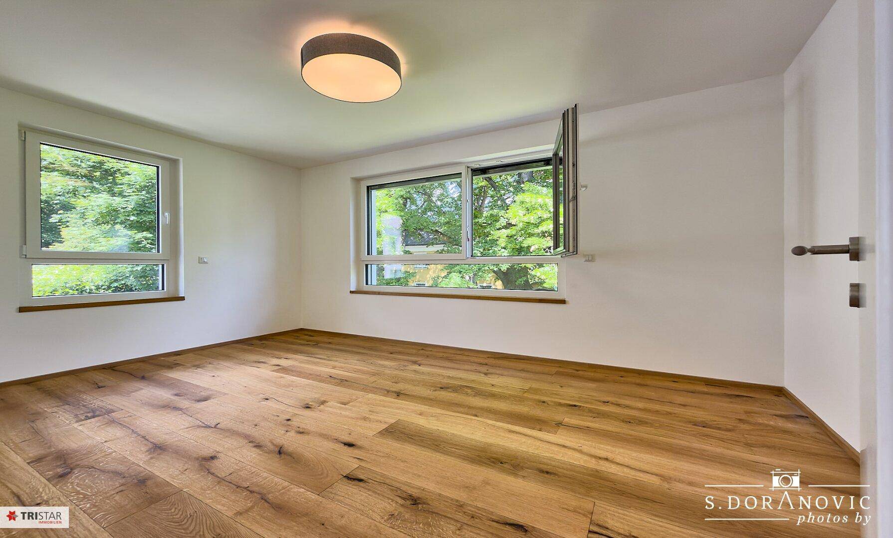 Geräumiges Schlafzimmer mit Holzboden und zwei Fenstern, die viel Tageslicht hereinlassen.