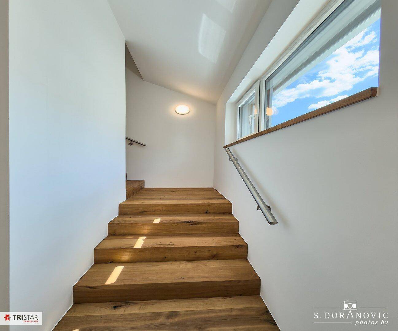 Helles Treppenhaus mit Holzstufen und modernem Geländer, das natürliches Licht empfängt.