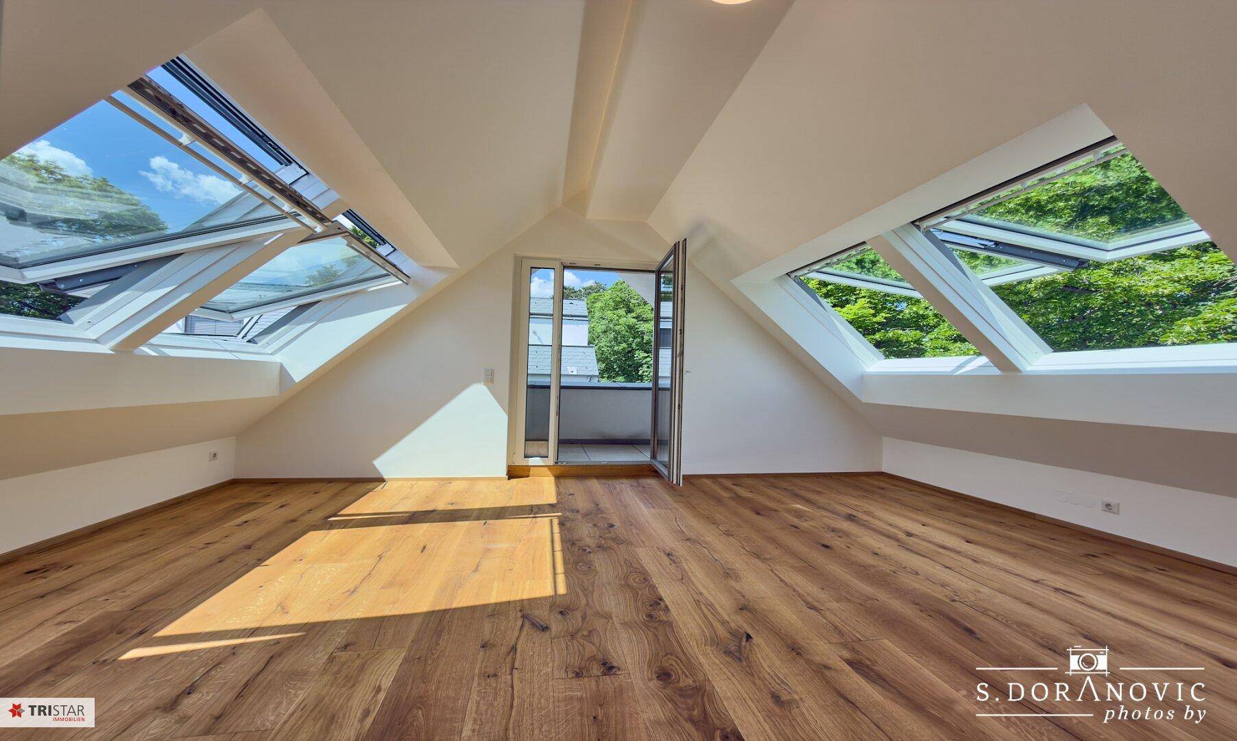 Helles Dachzimmer mit Holzboden, Schrägdach und großen Dachfenstern mit Grünblick.