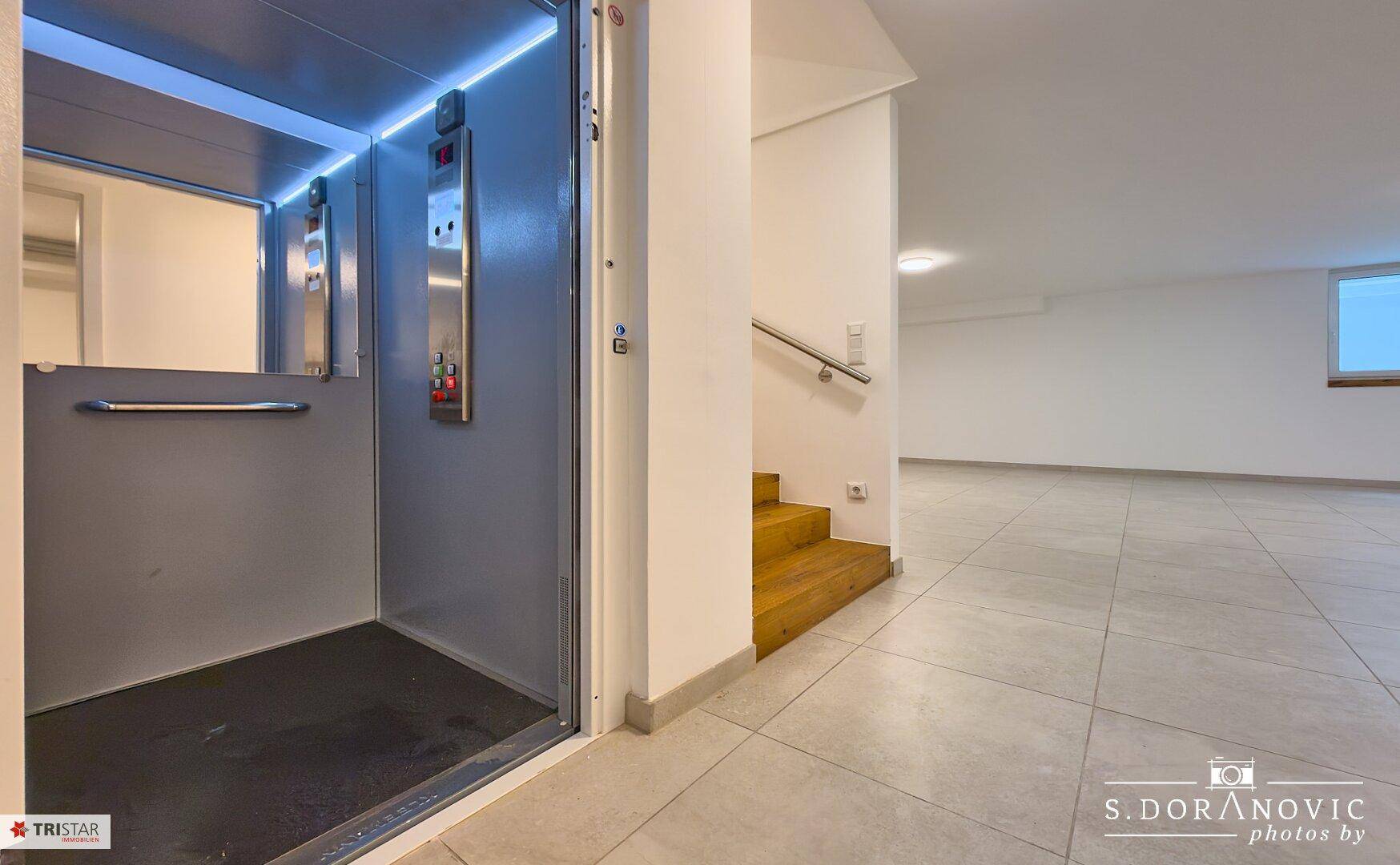 Moderner Aufzug mit geöffneter Tür und angrenzendem Treppenhaus mit Fliesenboden.