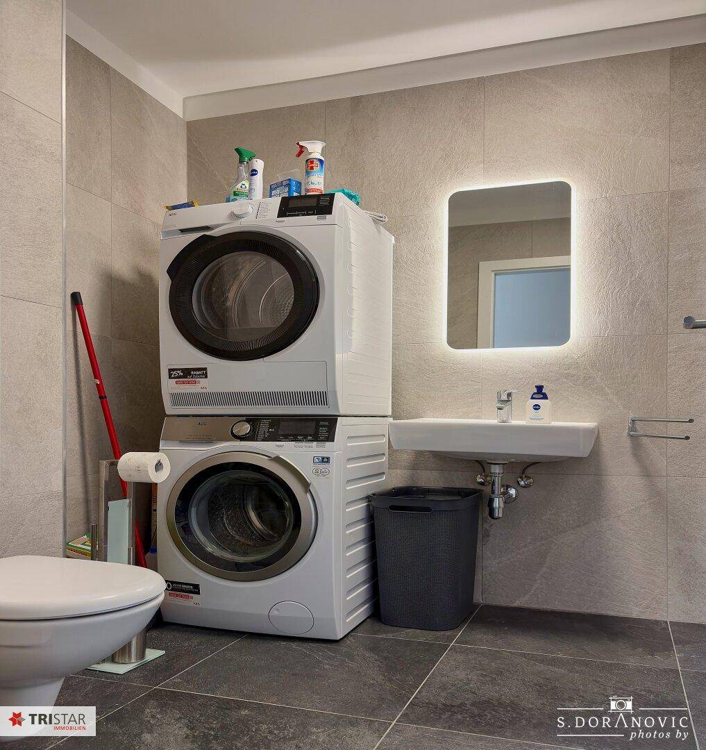 Modernes Badezimmer mit Waschmaschine, Trockner und beleuchtetem Spiegel.