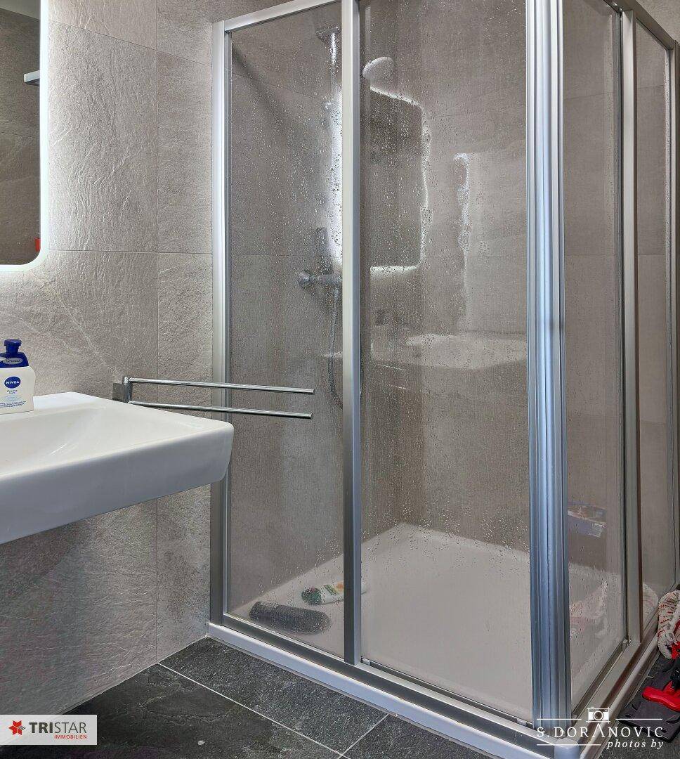Duschbereich in einem modernen Badezimmer mit grauen Fliesen und Glasduschkabine.