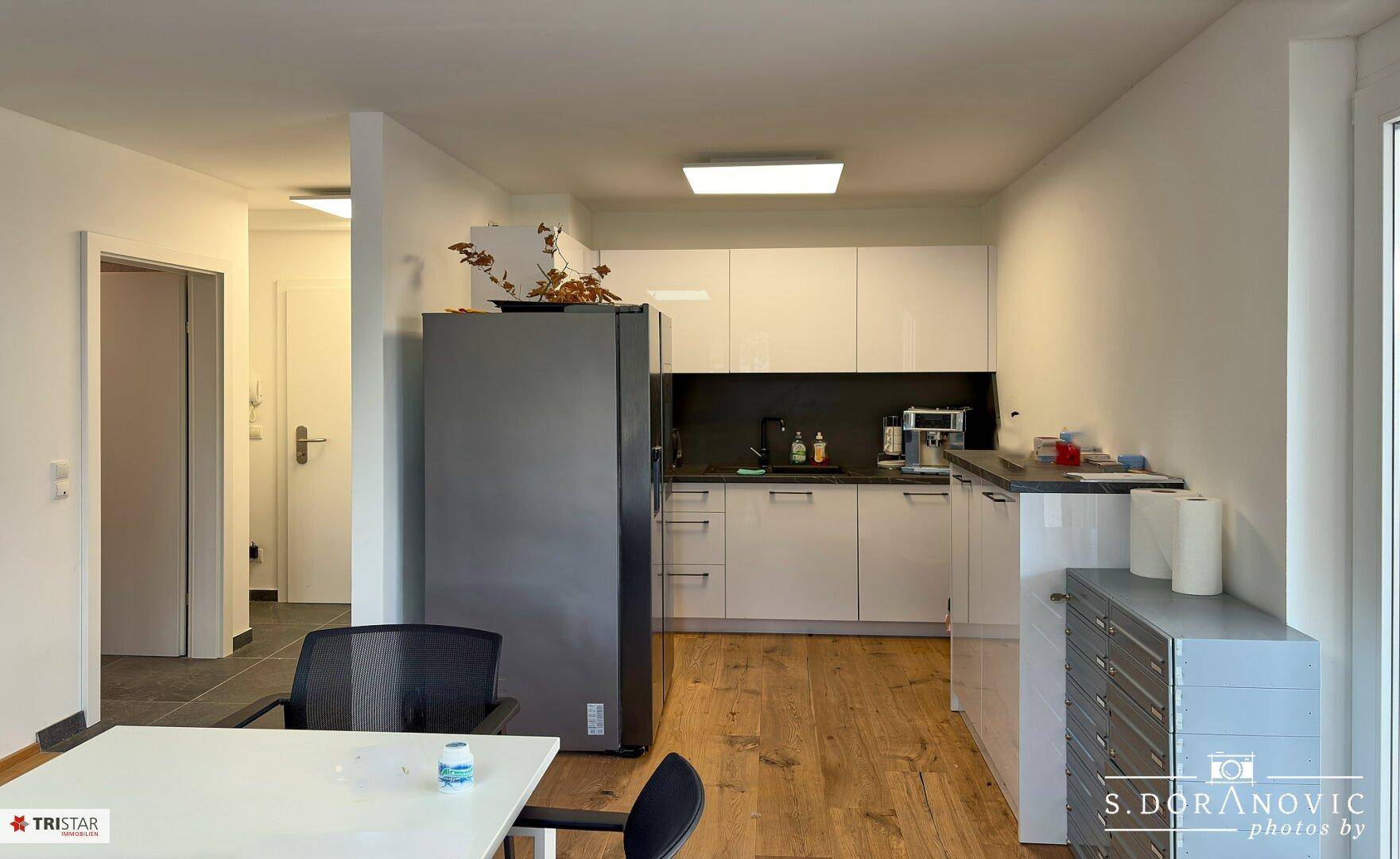 Moderne Küchenzeile in einem Büroraum mit Kühlschrank und weißen Schränken.