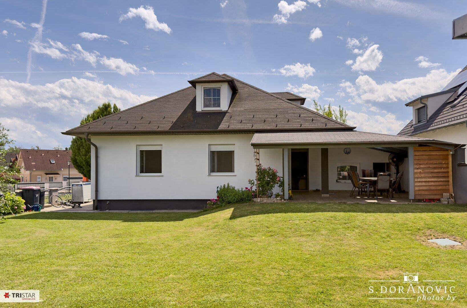 Rückseite des Hauses mit überdachter Terrasse und großem Gartenbereich unter blauem Himmel.