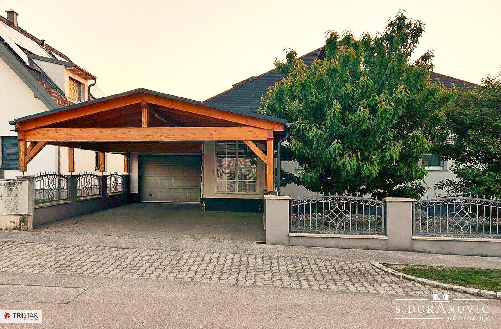 Holzcarport und Garage am Abend, beleuchtet und mit zwei geparkten Autos.