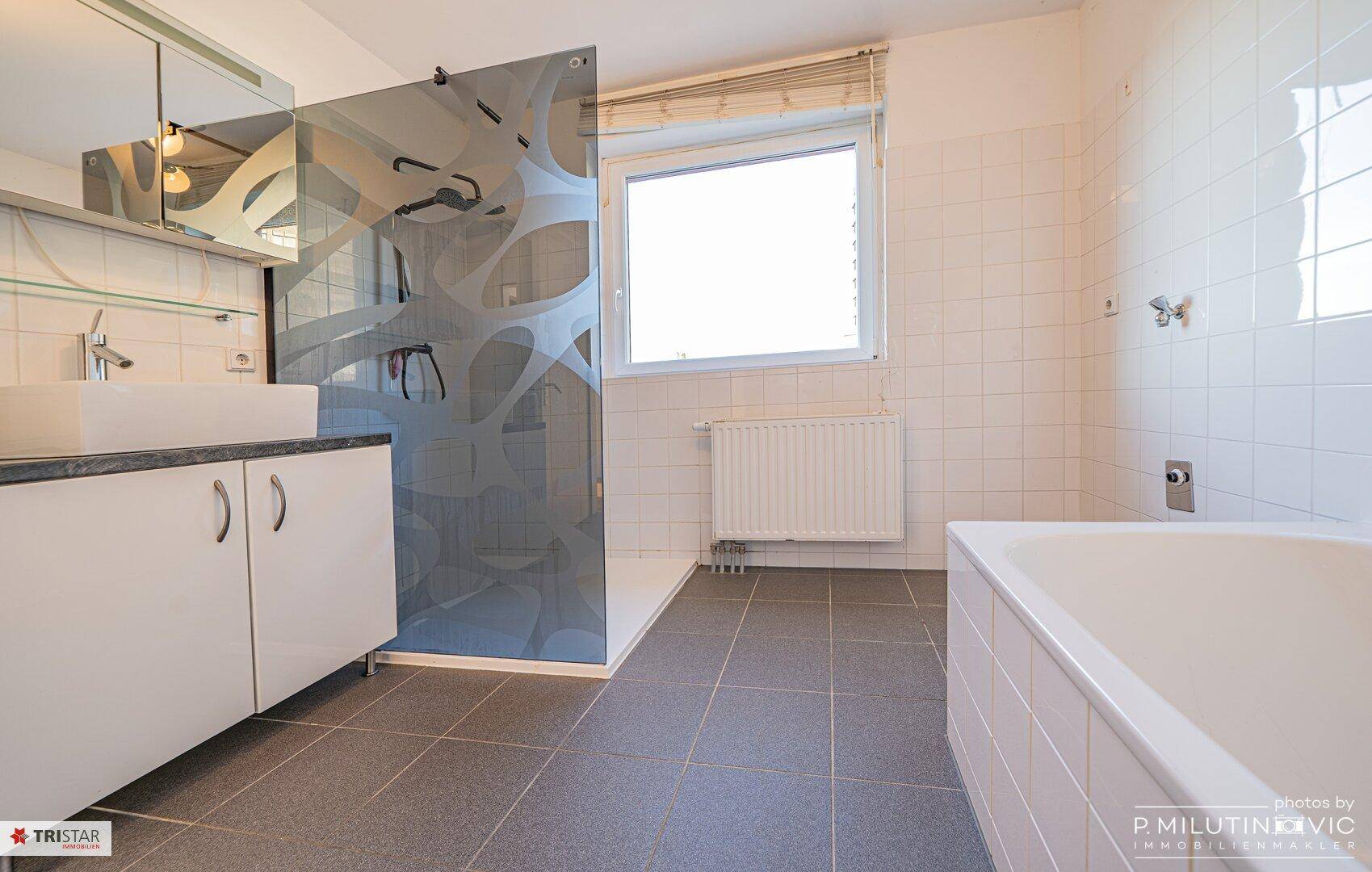 Helles Badezimmer mit Badewanne, Dusche, Einzelwaschbecken und Fenster.