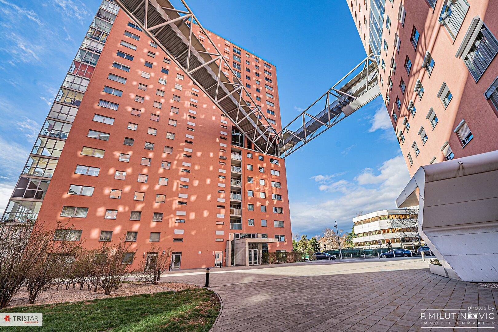 Zwei moderne Hochhäuser mit rötlicher Fassade, verbunden durch eine architektonische Brücke.